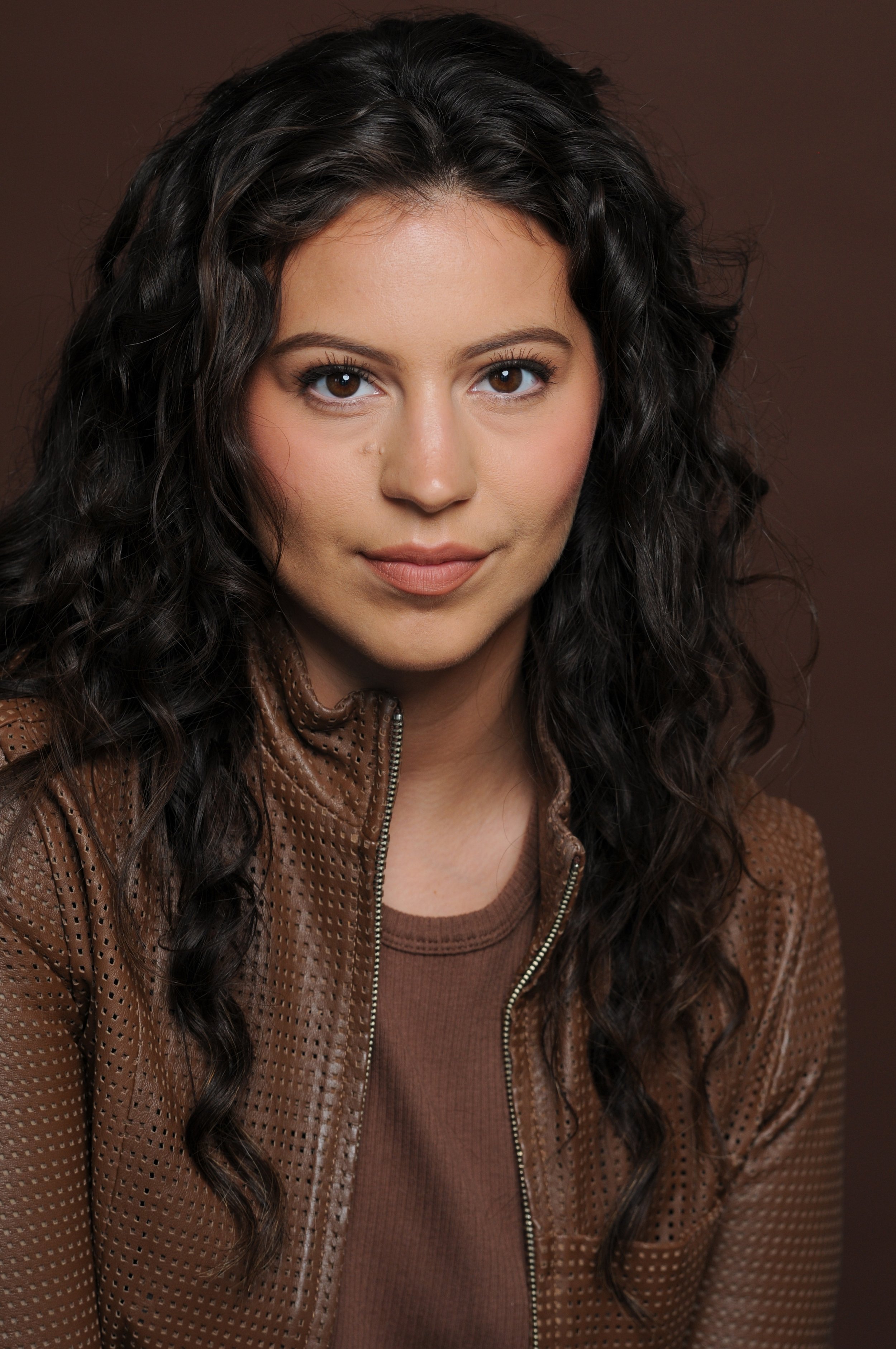 A woman with long, curly dark hair and brown eyes, wearing a brown leather jacket and a brown shirt, looking directly at the camera against a plain brown background.