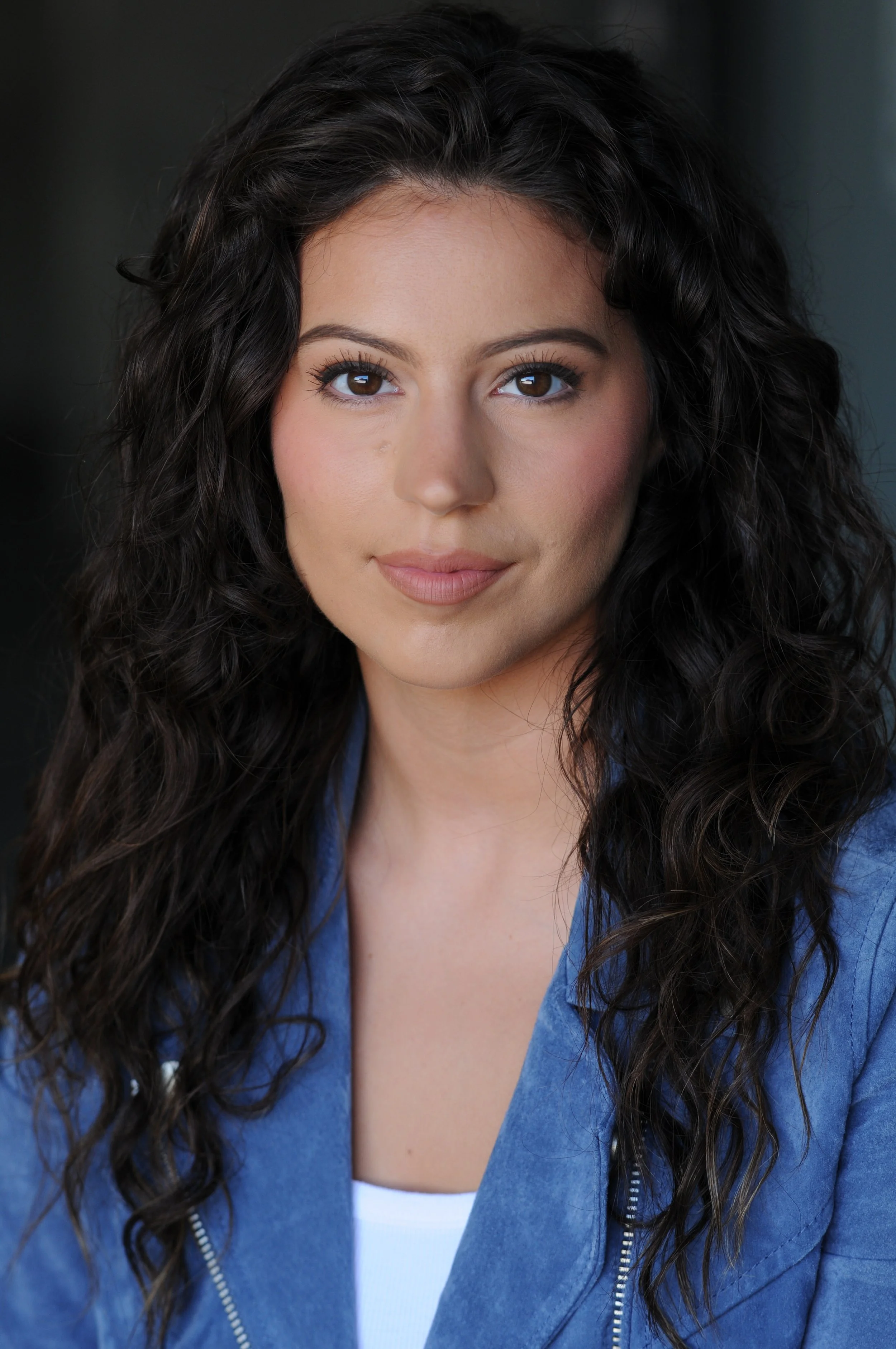 Close-up portrait of a young woman with long curly dark hair, wearing a blue jacket and a white top, looking directly at the camera with a slight smile.