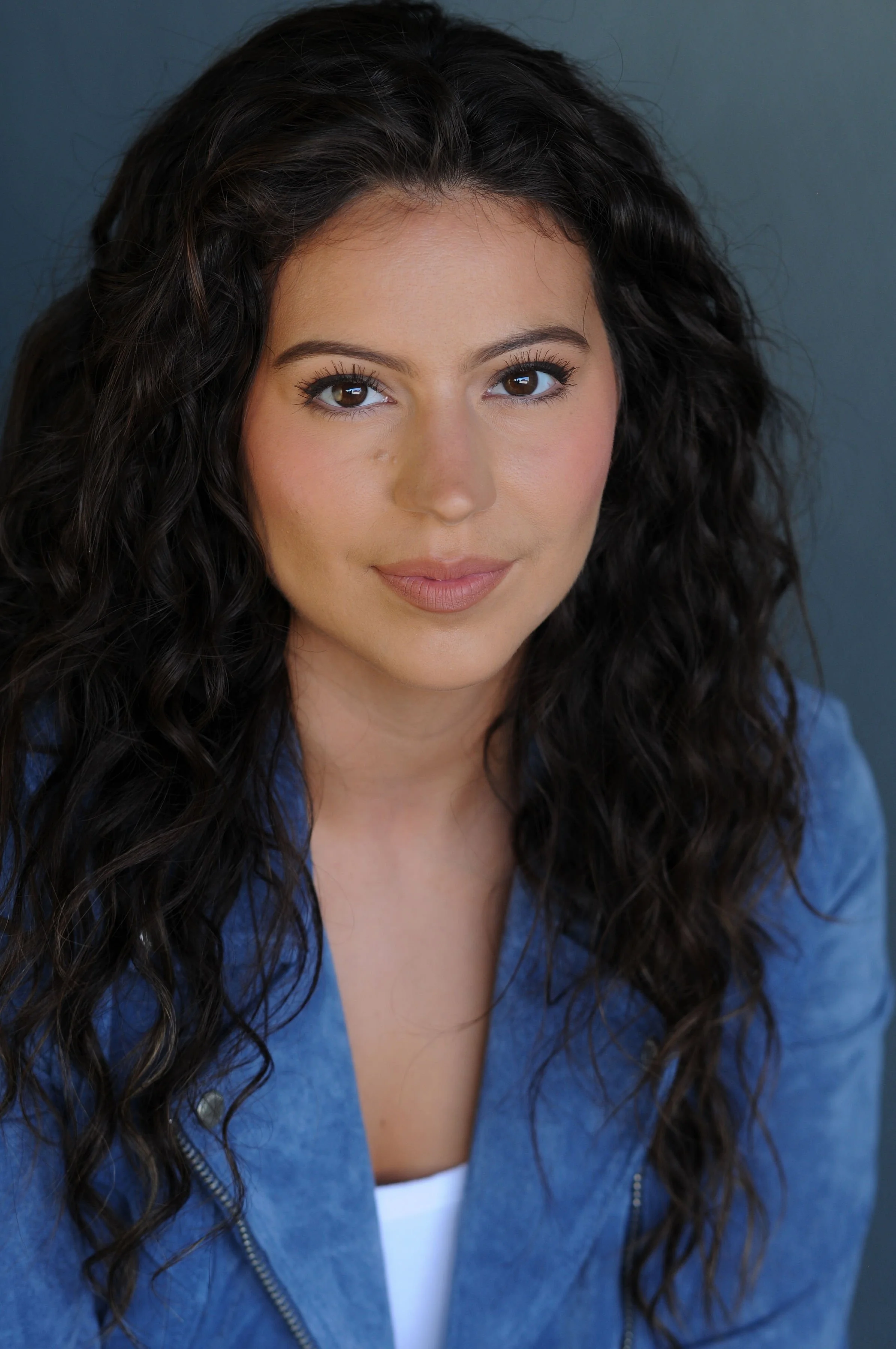 Portrait of a woman with long, curly dark hair wearing a blue jacket and a white top, looking at the camera with a slight smile.