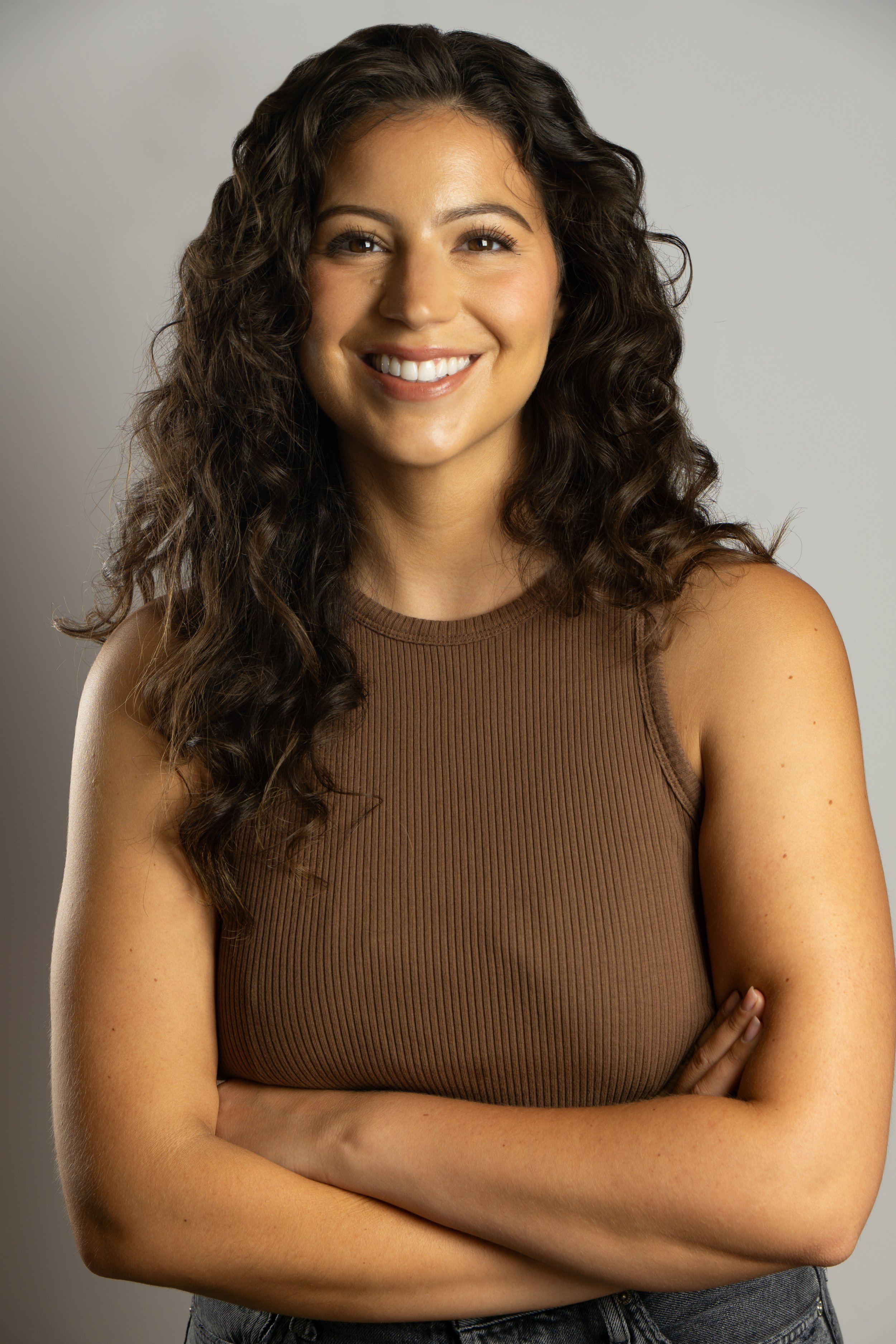 A woman with long curly brown hair smiling, wearing a sleeveless brown top and crossed arms, standing against a plain gray background.