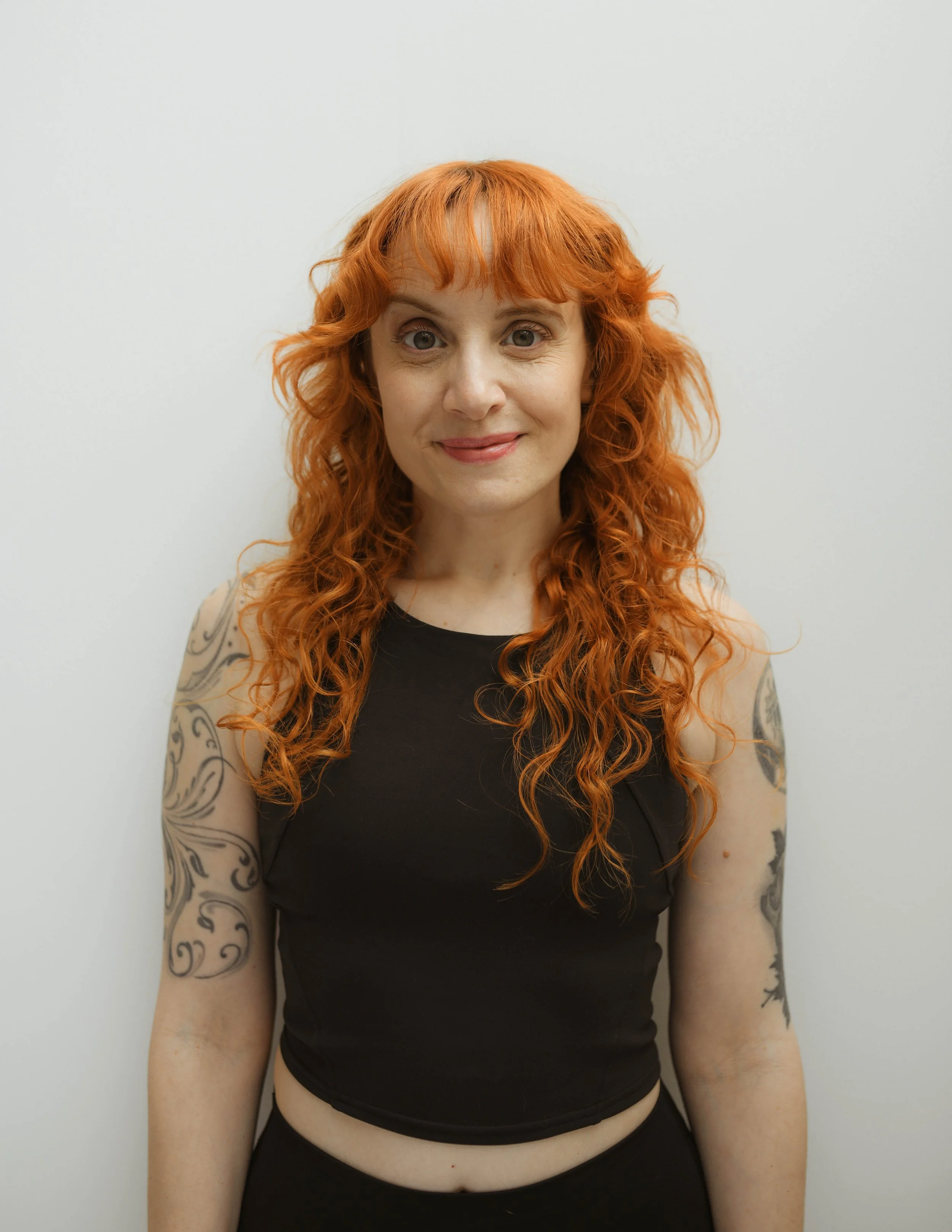 A woman with long dark wavy hair, wearing a black sleeveless top with a high neck, standing in front of a plain white wall, smiling at the camera.
