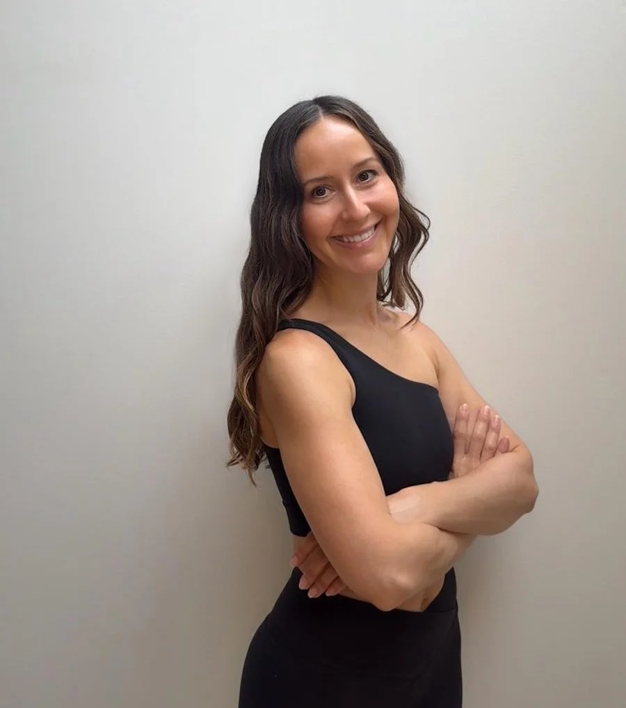 A woman with dark wavy hair smiling, wearing a black top, in front of a white wall.