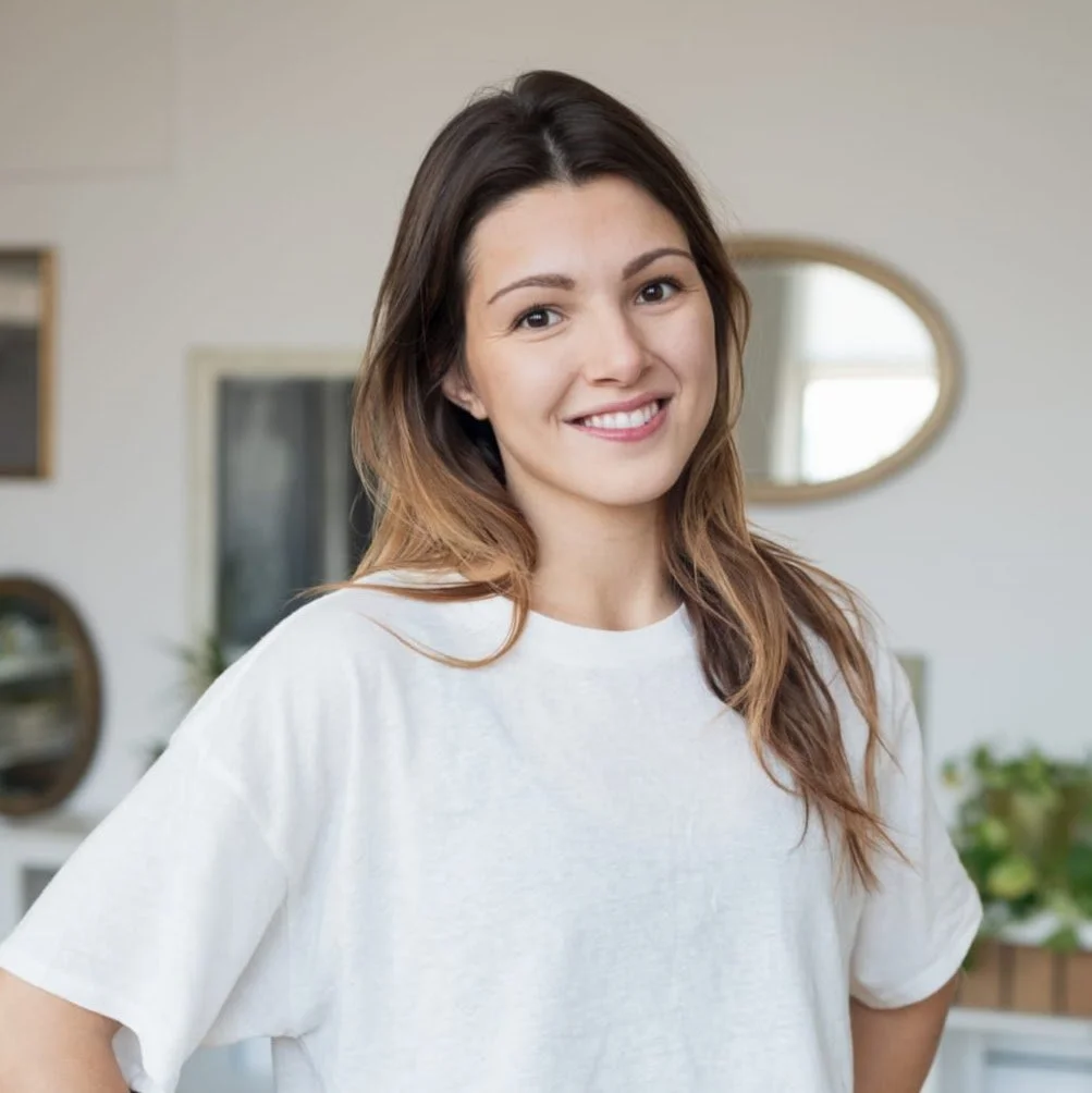 A woman with long dark wavy hair, wearing a black sleeveless top with a high neck, standing in front of a plain white wall, smiling at the camera.