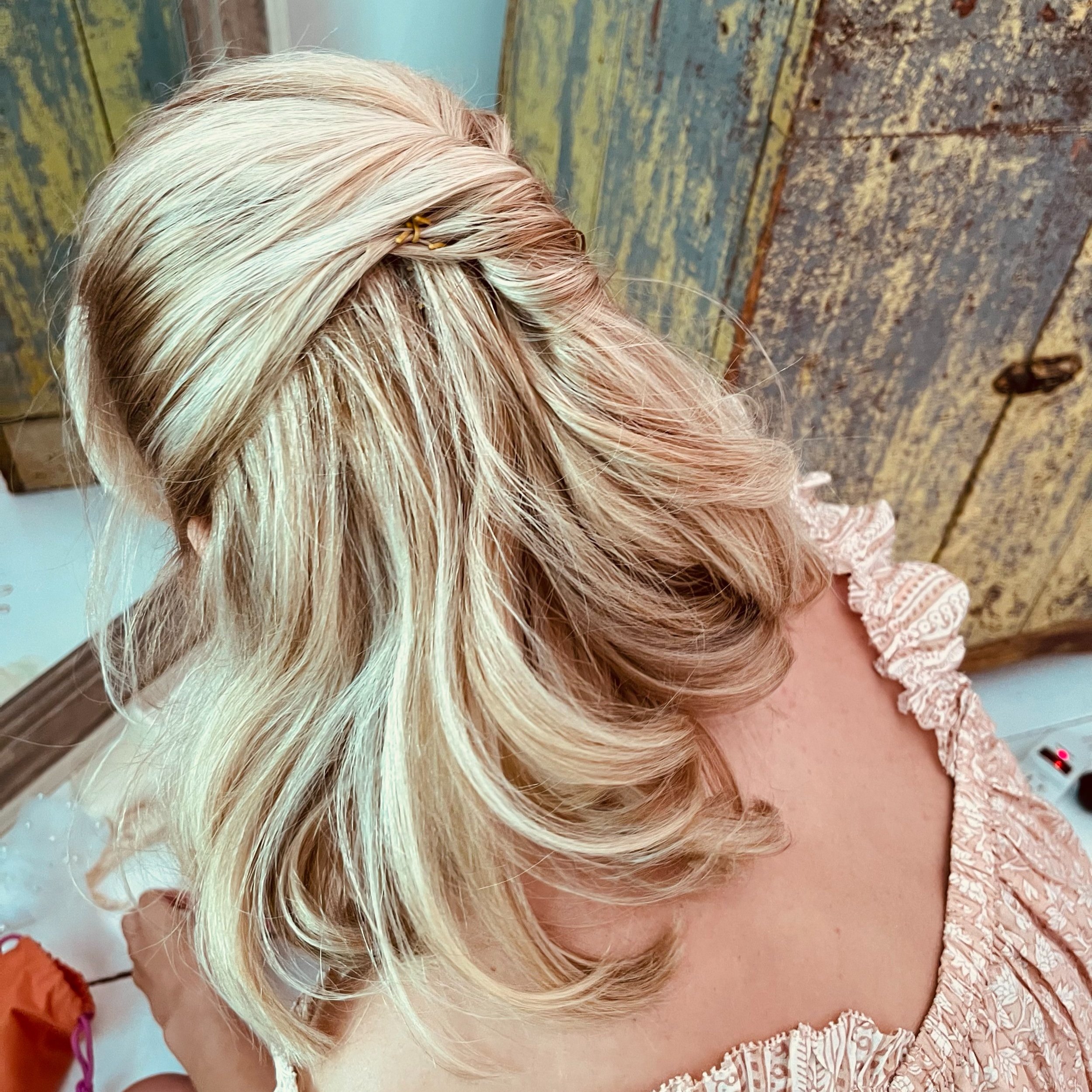 Blonde woman with styled hair, wearing a pink ruffled top, near a rustic wooden background.
