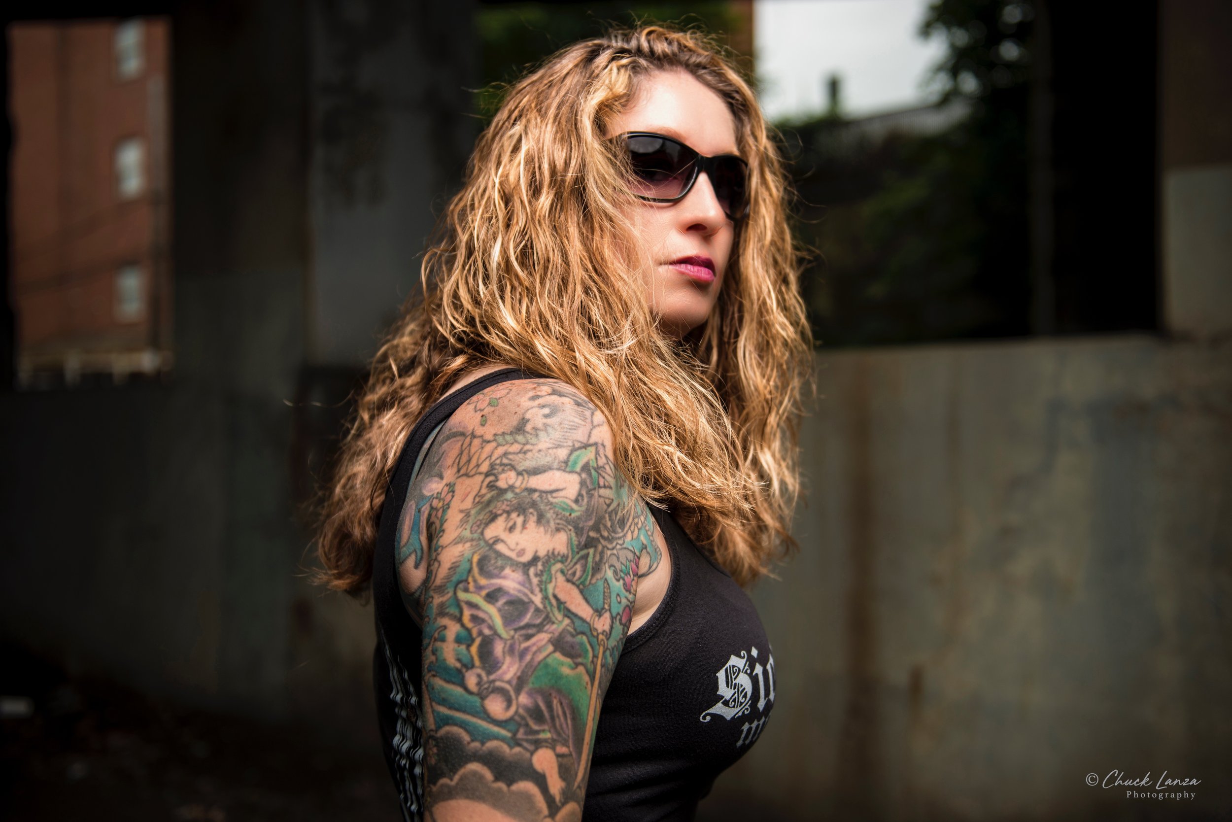 A woman with long curly blonde hair, wearing dark sunglasses, a black sleeveless top, and showing colorful tattoos on her upper arm, stands outdoors in an urban environment.