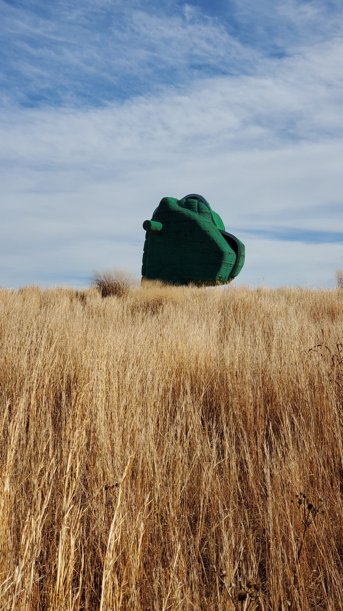 A large, green, tank-shaped structure positioned in a grassy field under a blue sky with scattered clouds.