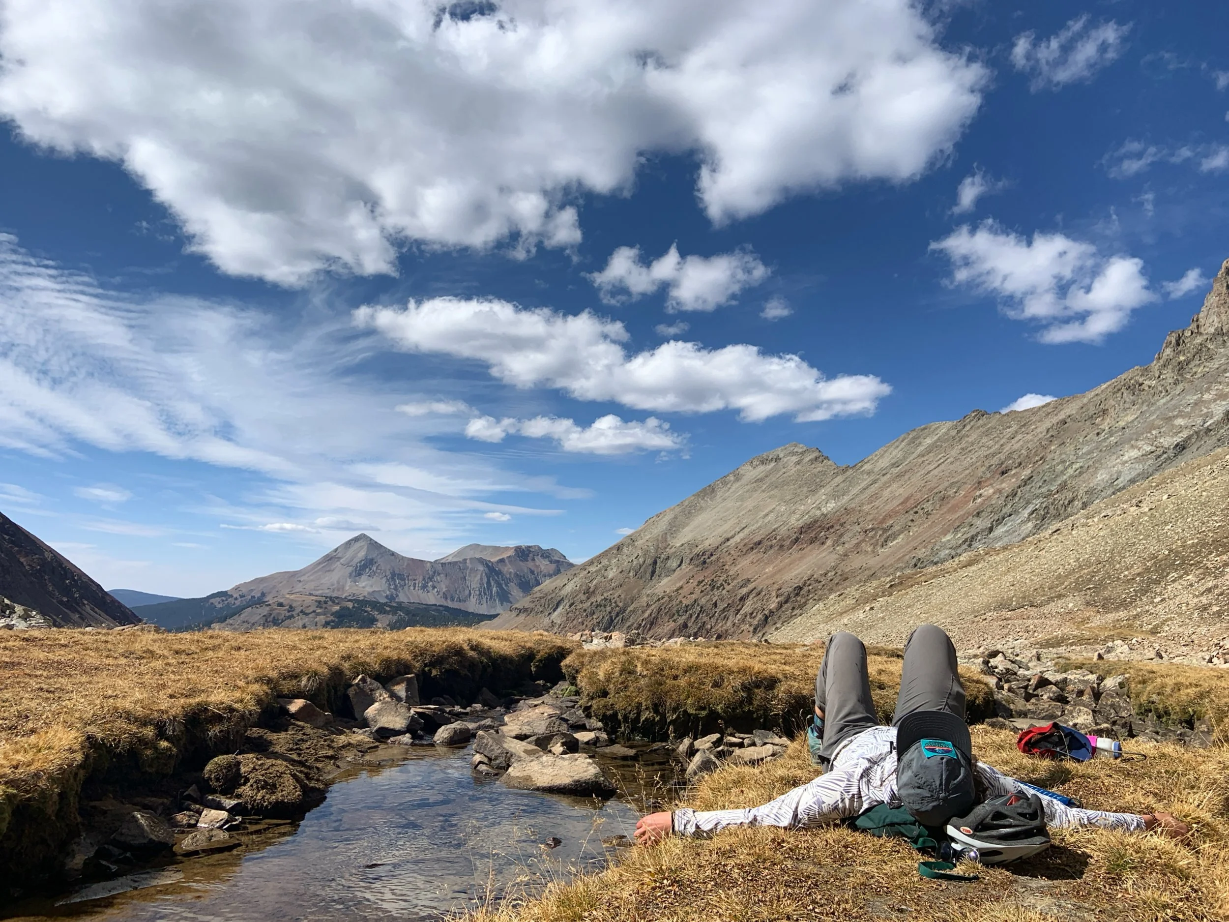 A person lying on grass near a small stream in a mountainous landscape with partly cloudy skies.