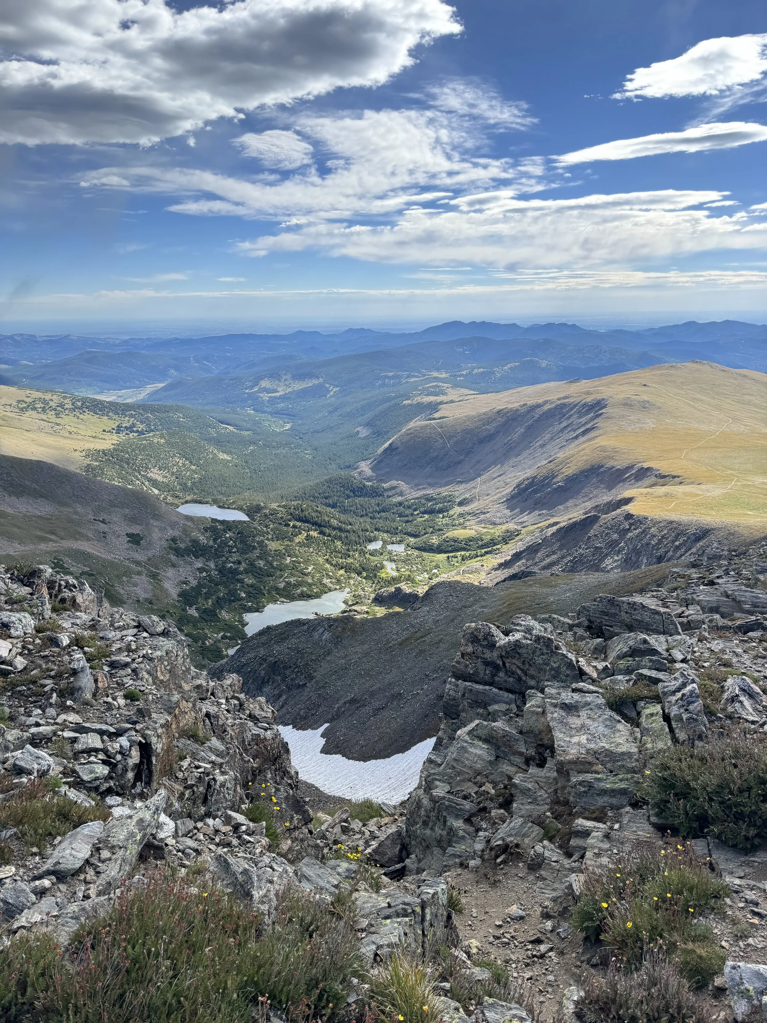 Mountain landscape with rocky foreground, lakes, rolling hills, and a partly cloudy sky.