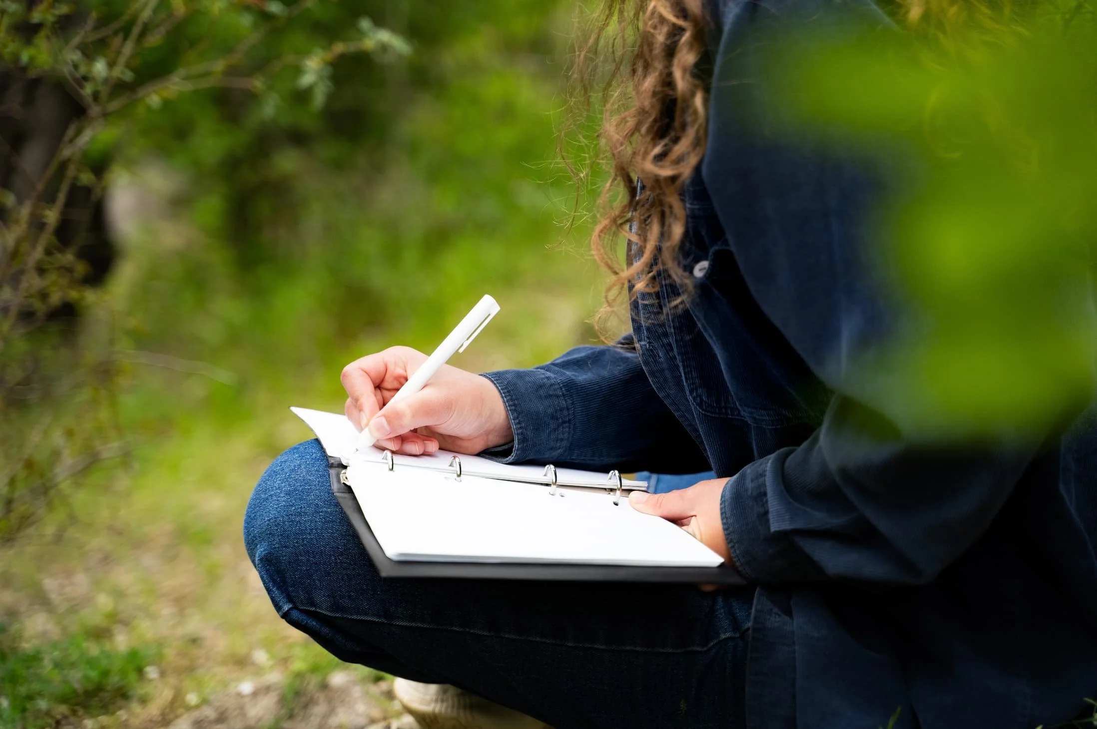 Person crouching outdoors, writing in a white notebook with a pen, surrounded by green foliage.