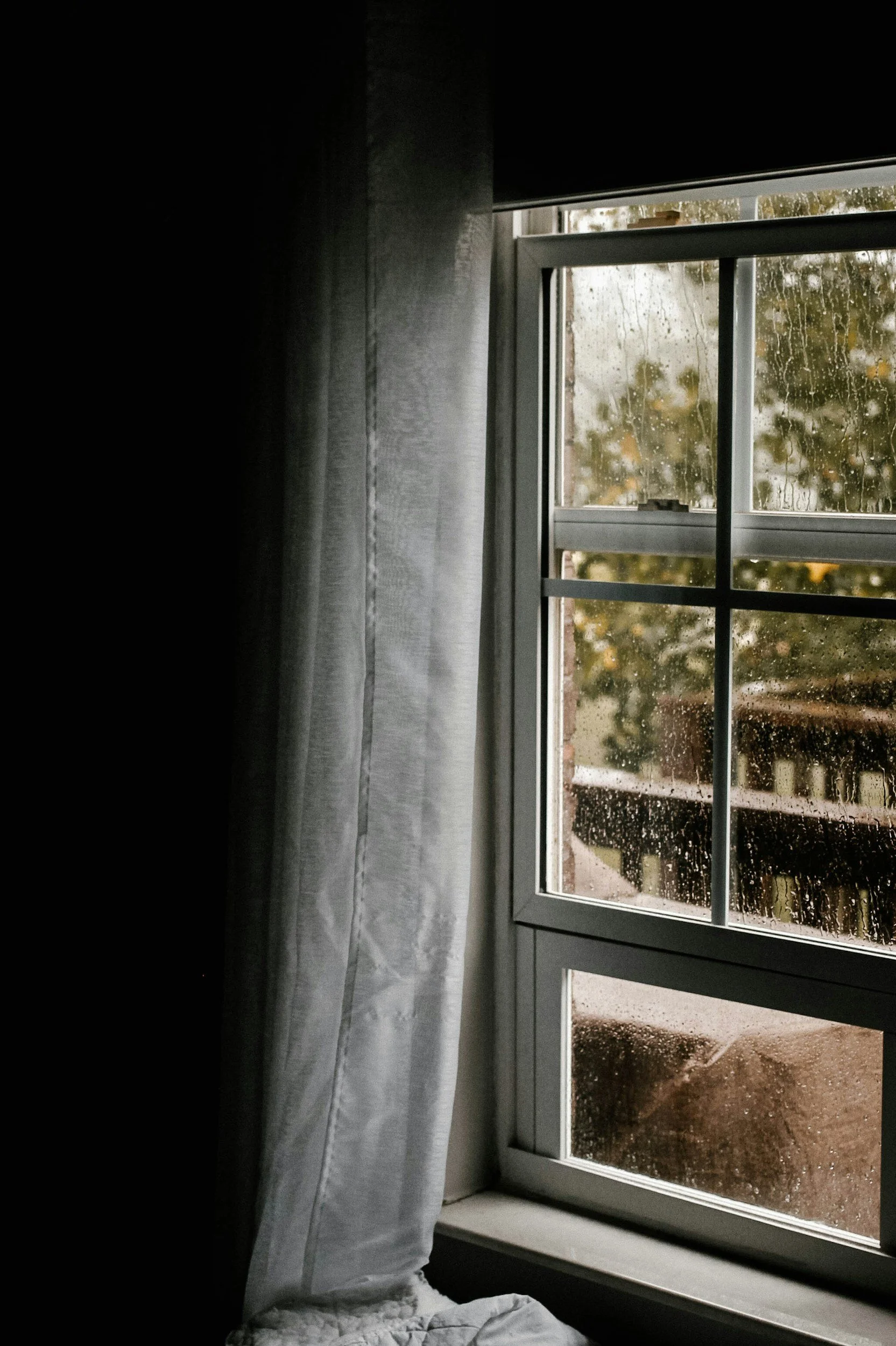 A window with raindrops on the glass and a partially drawn white curtain inside a room, showing an outdoor scene with trees and a wooden fence on a rainy day.