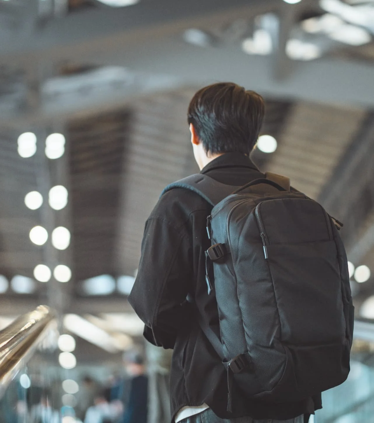 Person with dark hair wearing a black jacket and carrying a black backpack standing in an airport terminal.