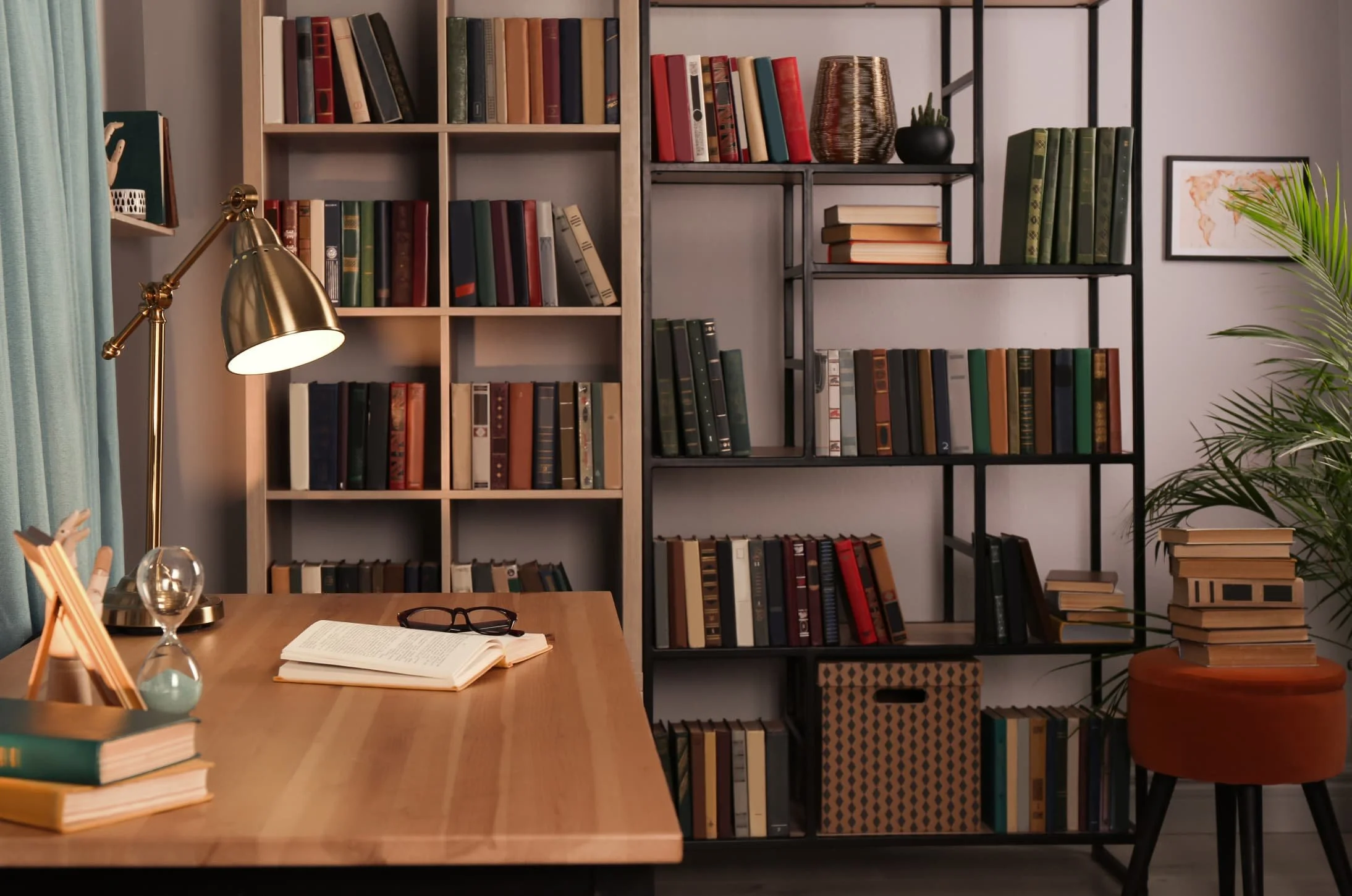 A cozy office or study space with a wooden desk, bookshelves filled with books, a gold desk lamp, an open book, eyeglasses, a glass paperweight, and a potted plant. The background includes a gray wall with framed artwork and a green curtain partially visible on the left.