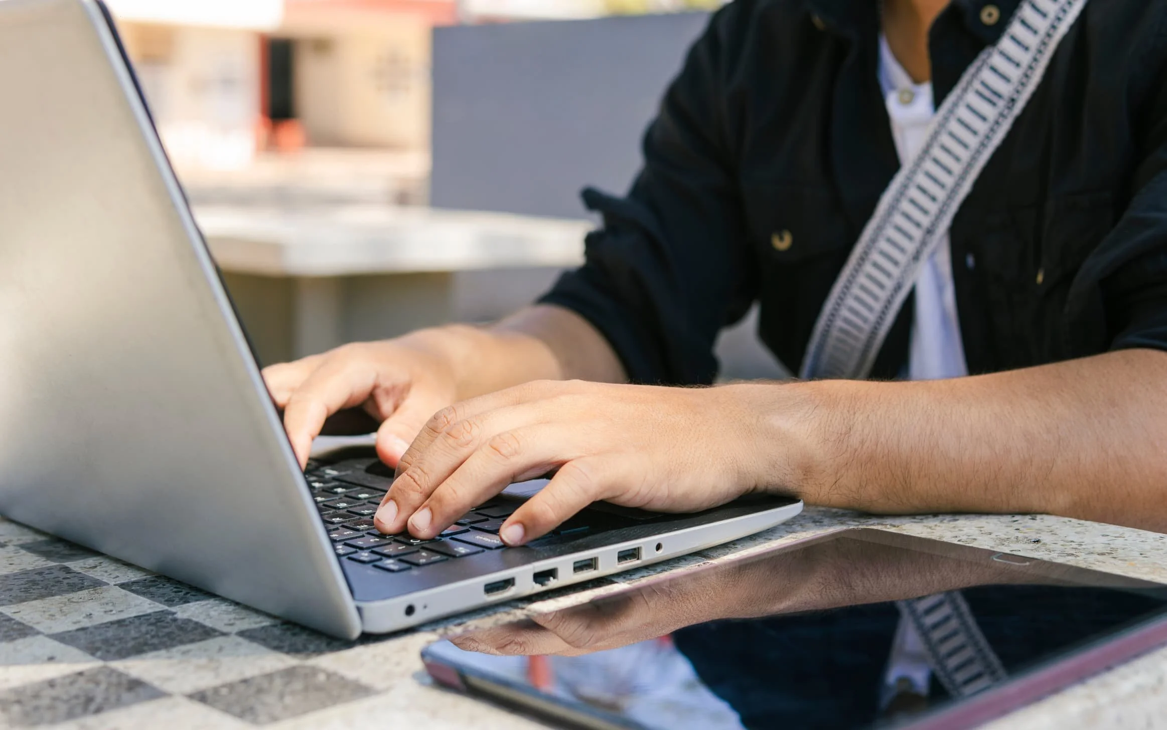 A person typing on a silver laptop with a black keyboard, with a smartphone placed on a checkered granite table next to the laptop.