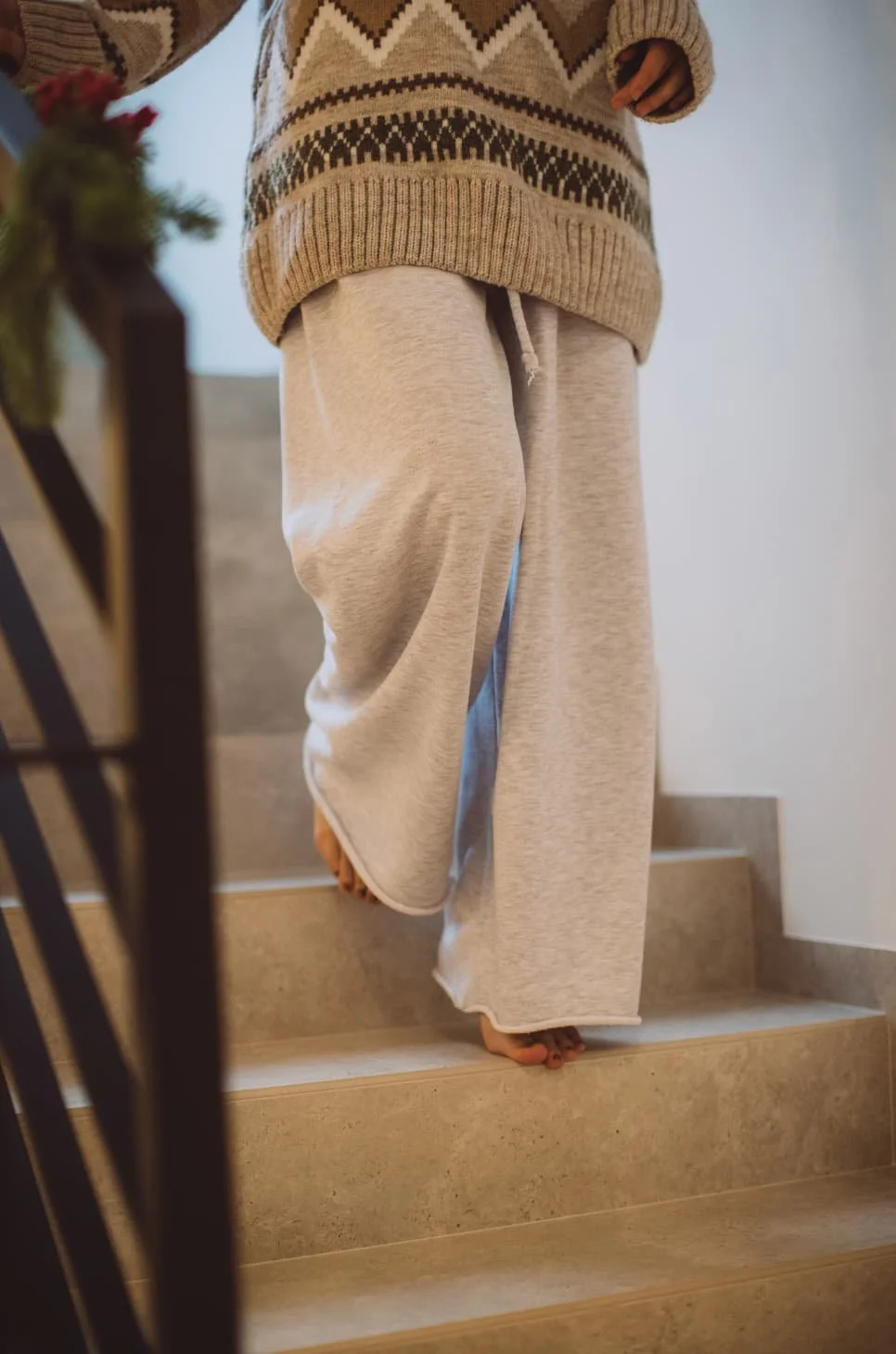 Person walking down stairs wearing a patterned sweater and loose light-colored sweatpants, with bare feet.