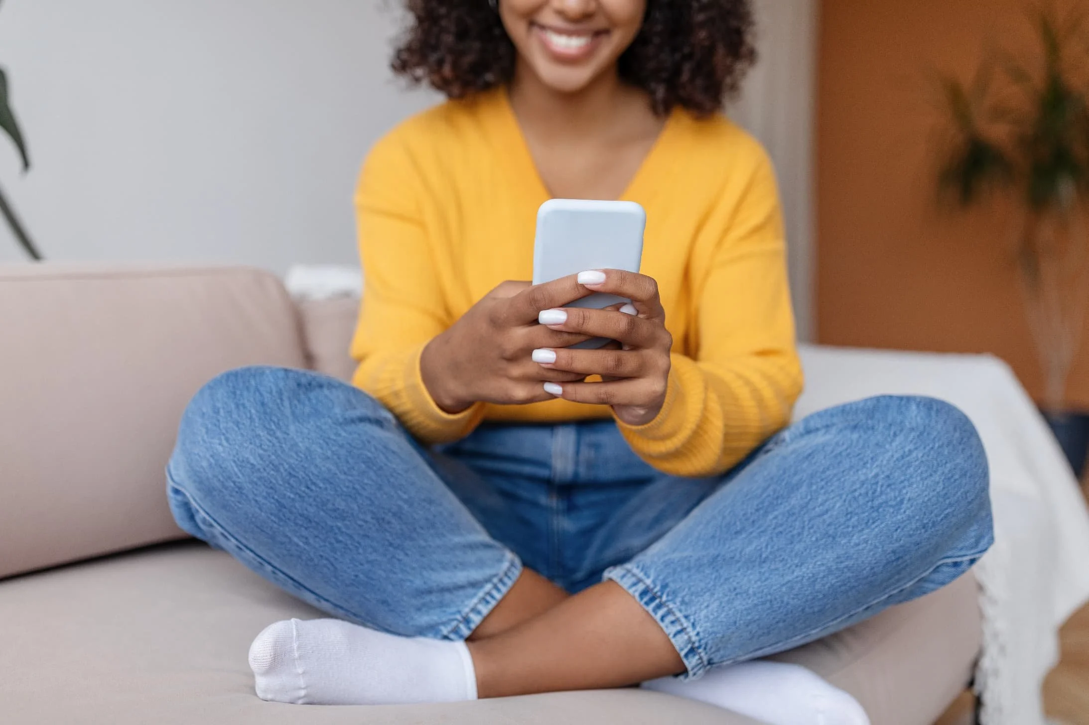 A woman sitting cross-legged on a beige sofa, wearing a yellow sweater and blue jeans, looking at her smartphone and smiling.
