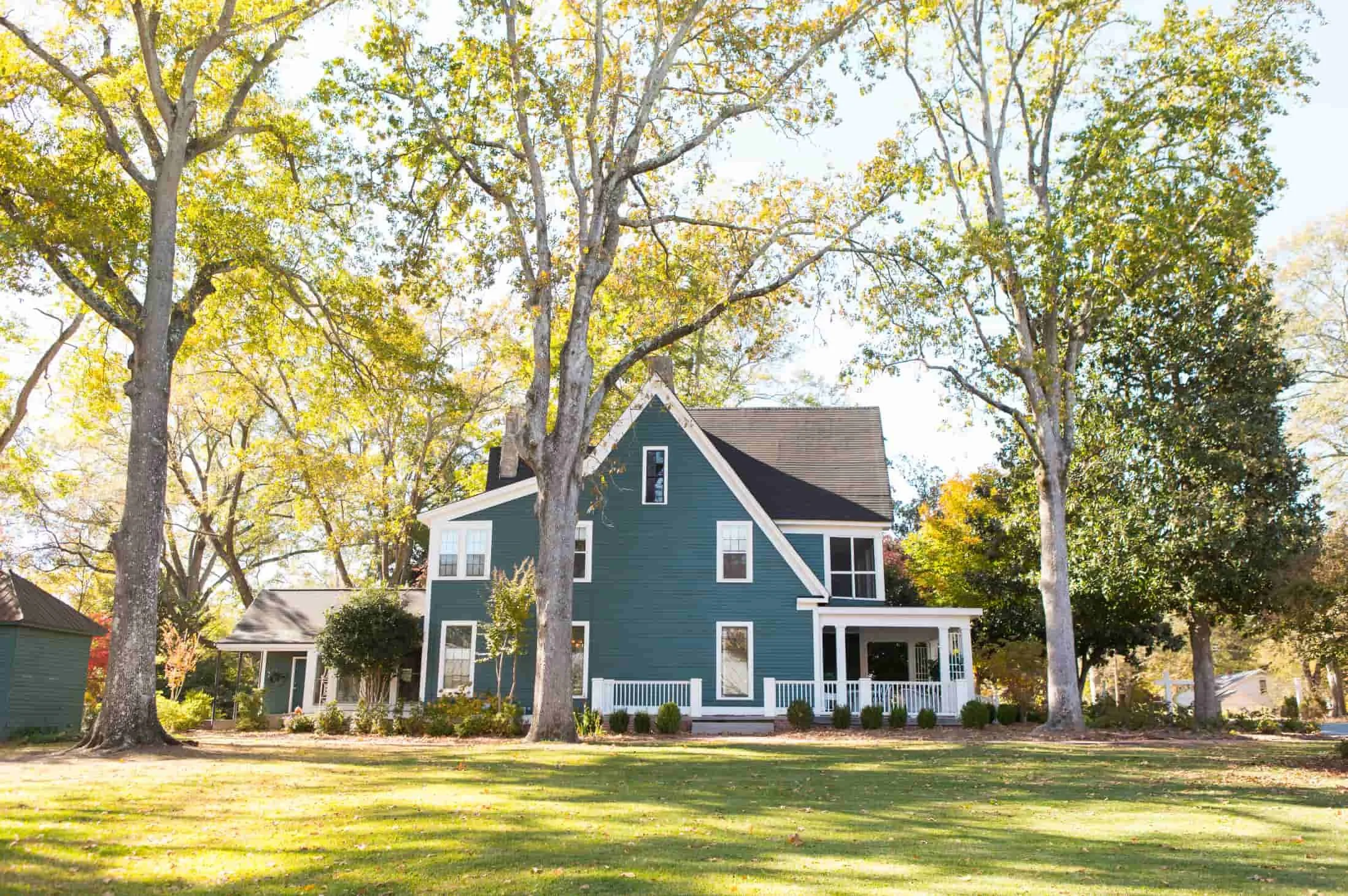A large blue house with white trim and a porch, surrounded by tall trees with green and yellow leaves, on a sunny day.