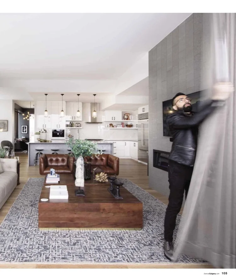 Man pulling back a curtain in a modern, well-lit living room with open kitchen, brown leather sofas, a wooden coffee table, and a patterned rug.
