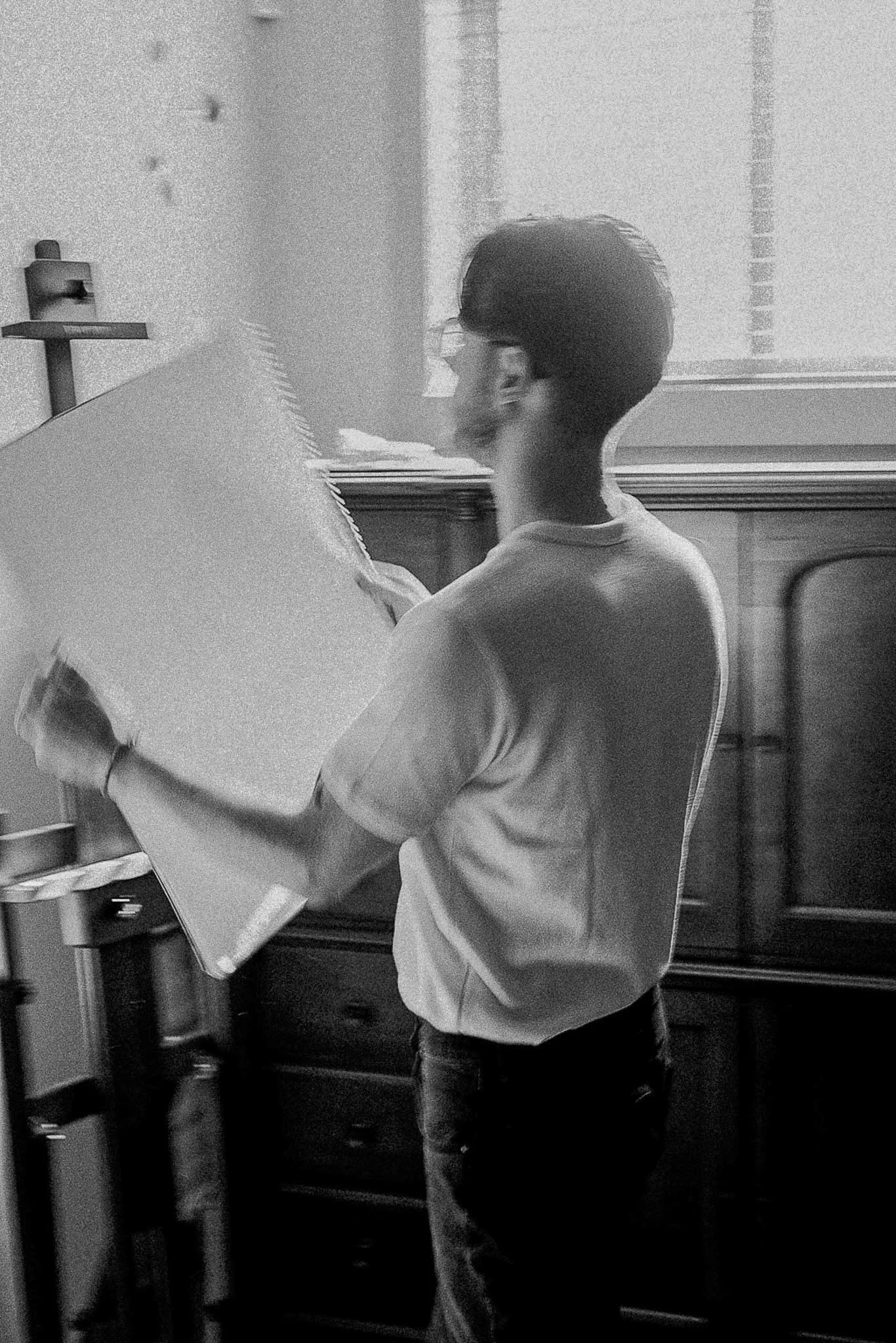 A young person with short dark hair and glasses standing indoors, holding and looking at an open notebook or sketchbook.