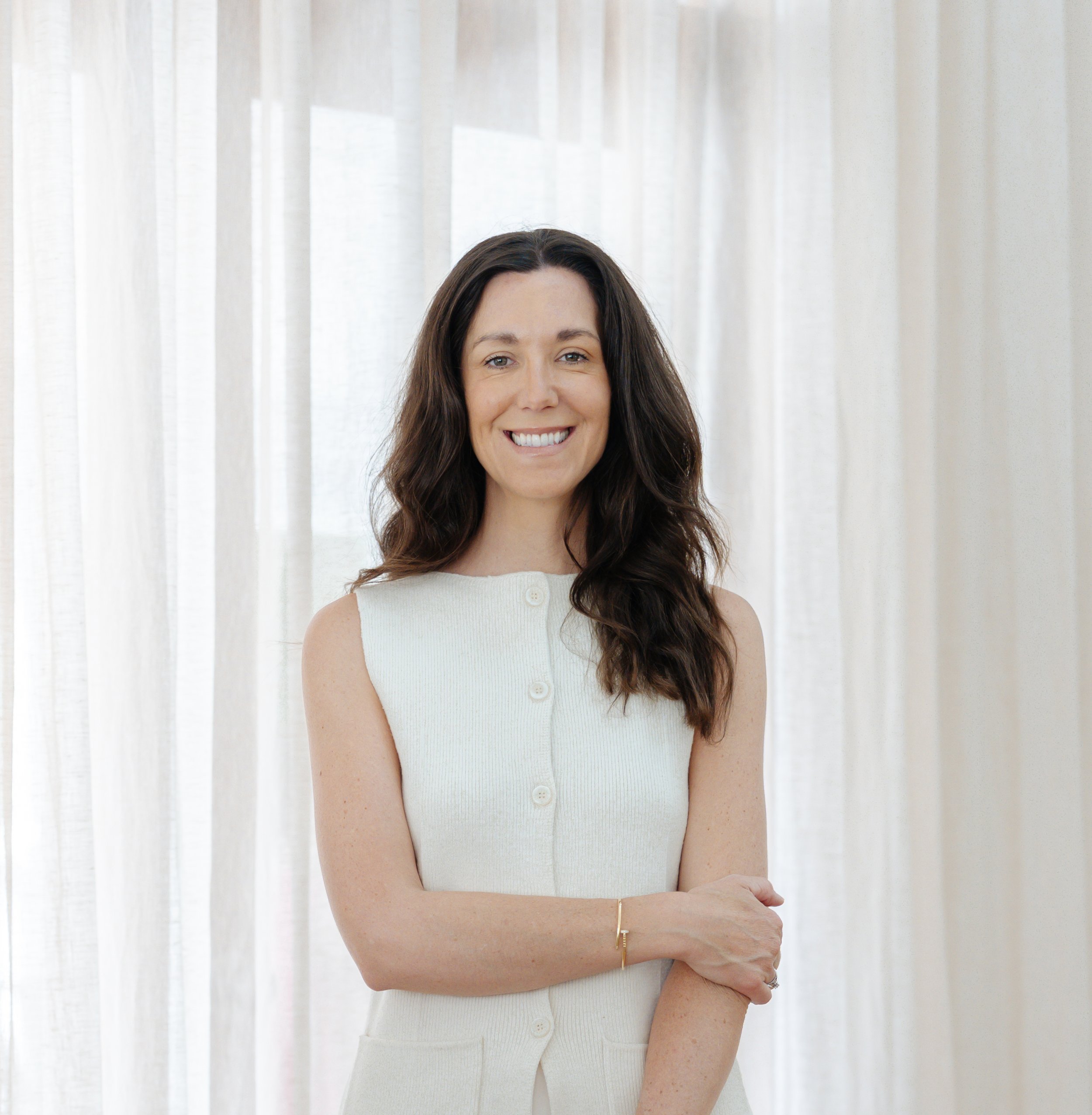 A woman with long dark brown hair smiling in front of sheer white curtains, wearing a sleeveless white knit top, with her arms crossed.