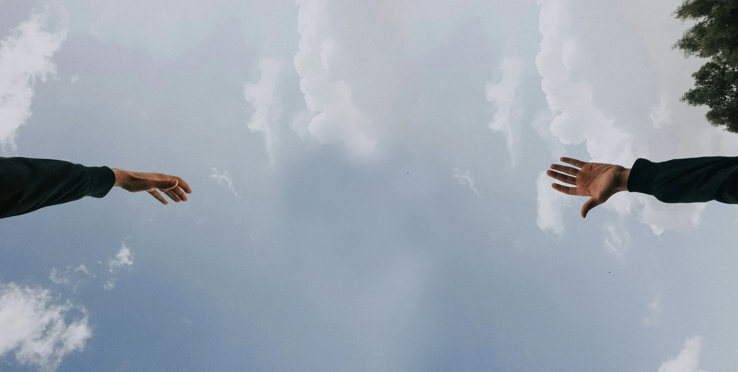 Two hands reaching towards each other against a cloudy sky, with trees on the right side.