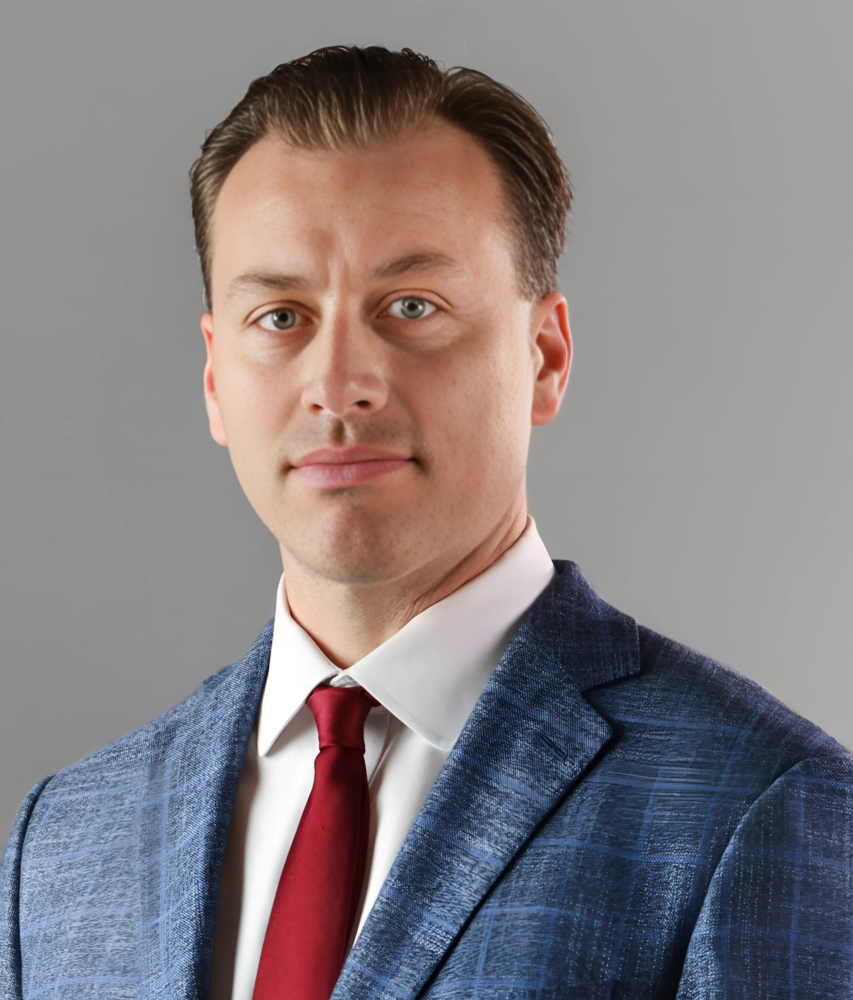 A man wearing a blue suit, white shirt, and red tie, looking directly at the camera with a neutral expression.
