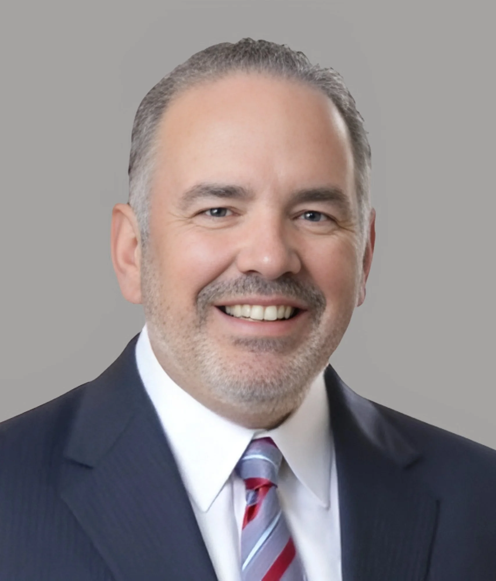 Headshot of a smiling man with short gray hair, beard, dressed in a dark suit, white shirt, and a striped tie, against a gray background.