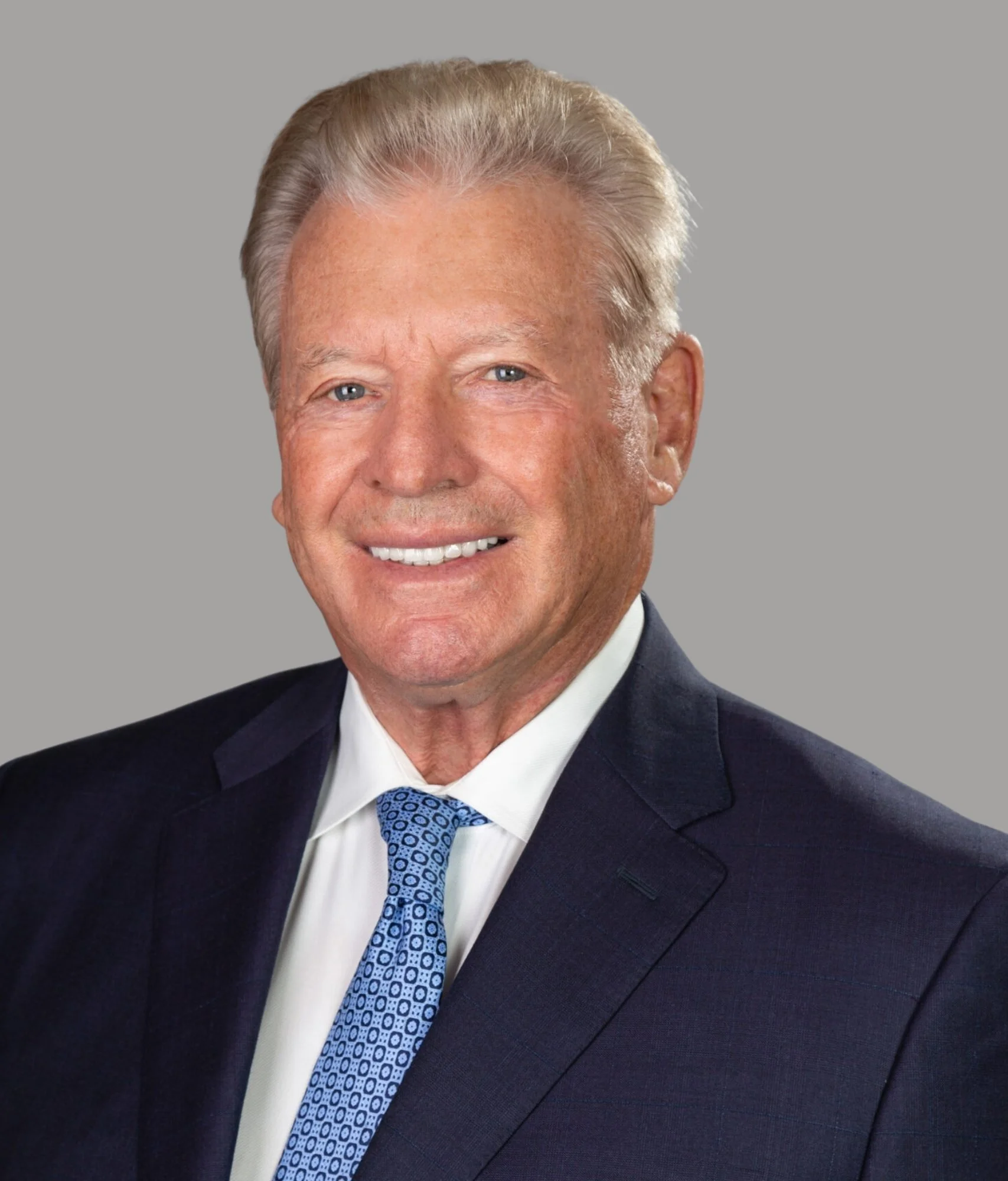 Portrait of an older man with gray hair wearing a dark suit, white shirt, and blue patterned tie, smiling against a gray background.