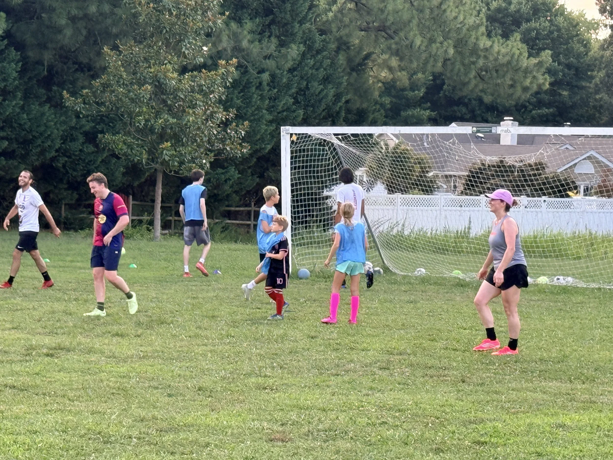 Children and adults playing soccer outdoors on a grassy field with a goalpost, trees, and houses in the background.