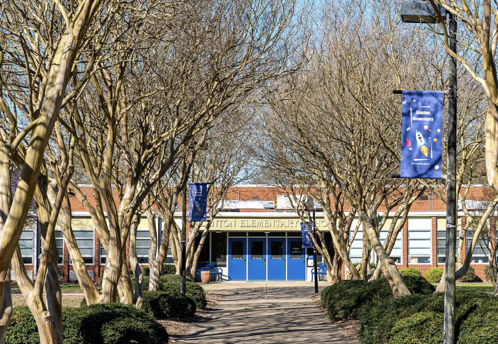 Entrance to Planton Elementary School with a walkway flanked by leafless trees and banners, blue doors under a curved roof, and brick building walls.