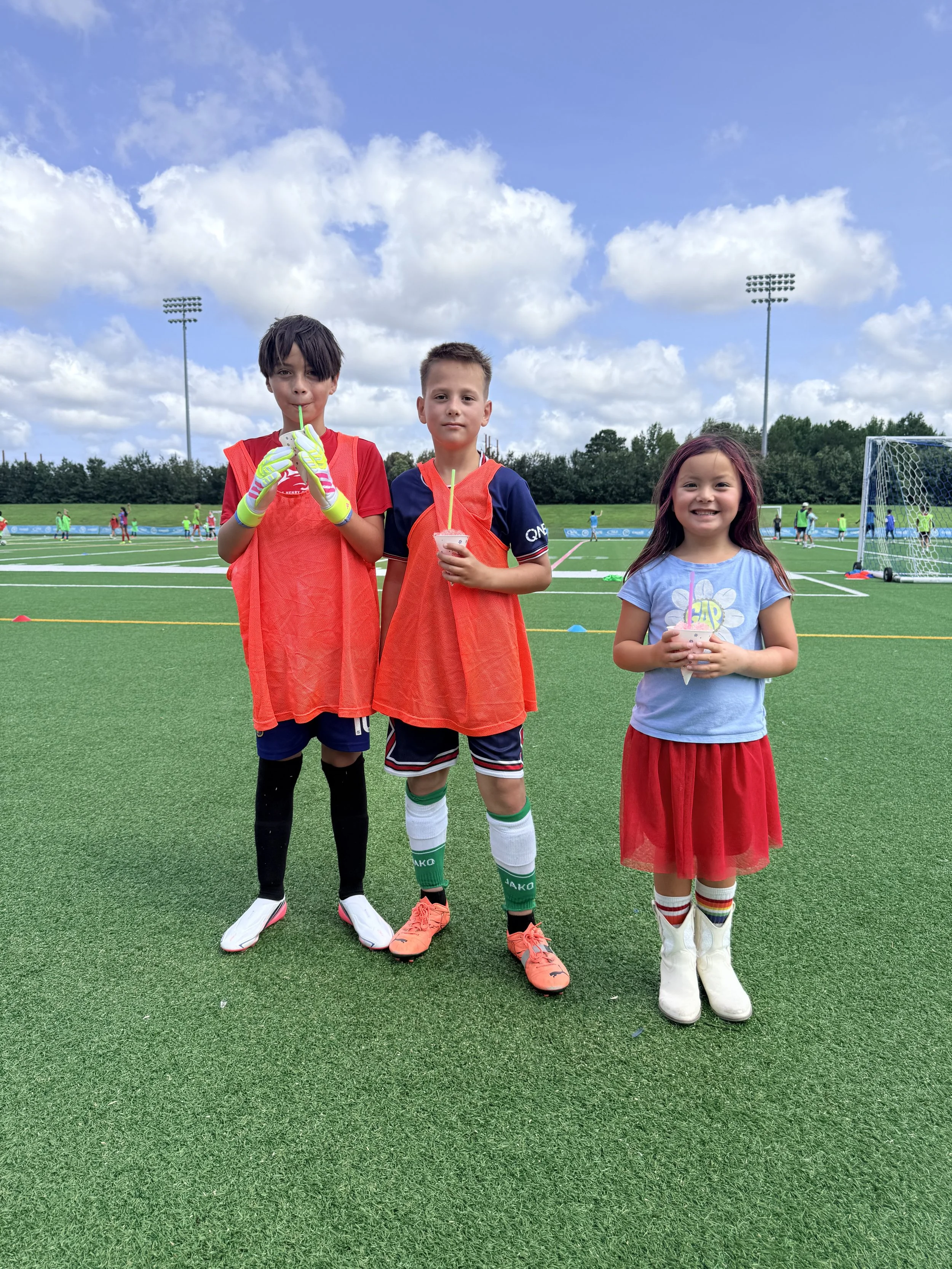 Three children standing on a soccer field, two boys wearing orange vests and a girl in a blue shirt and red skirt, holding drinks, with soccer goals and other children in the background under a cloudy sky.