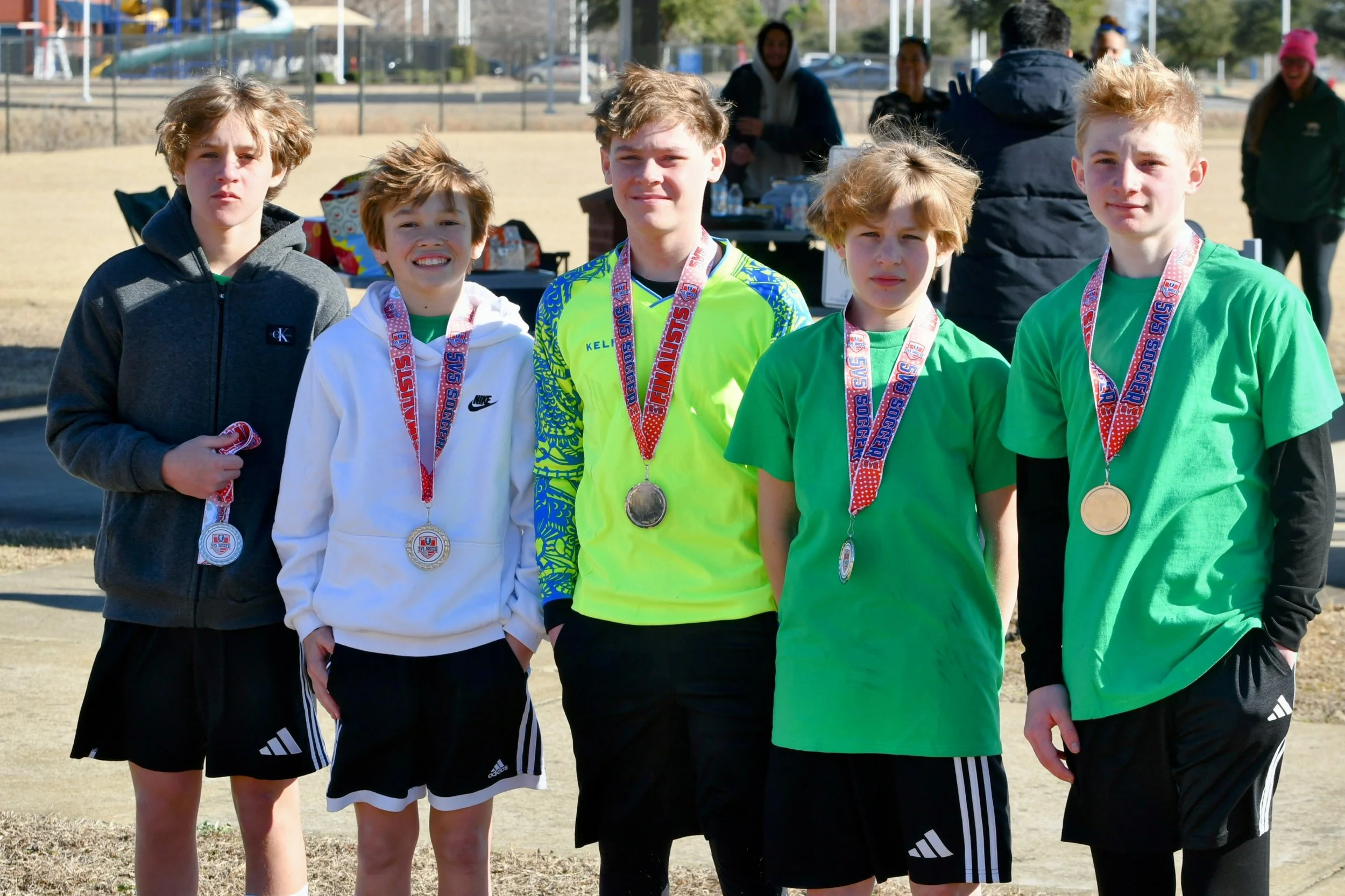 Five young boys standing outdoors wearing medals, some in sports attire, after a soccer event.