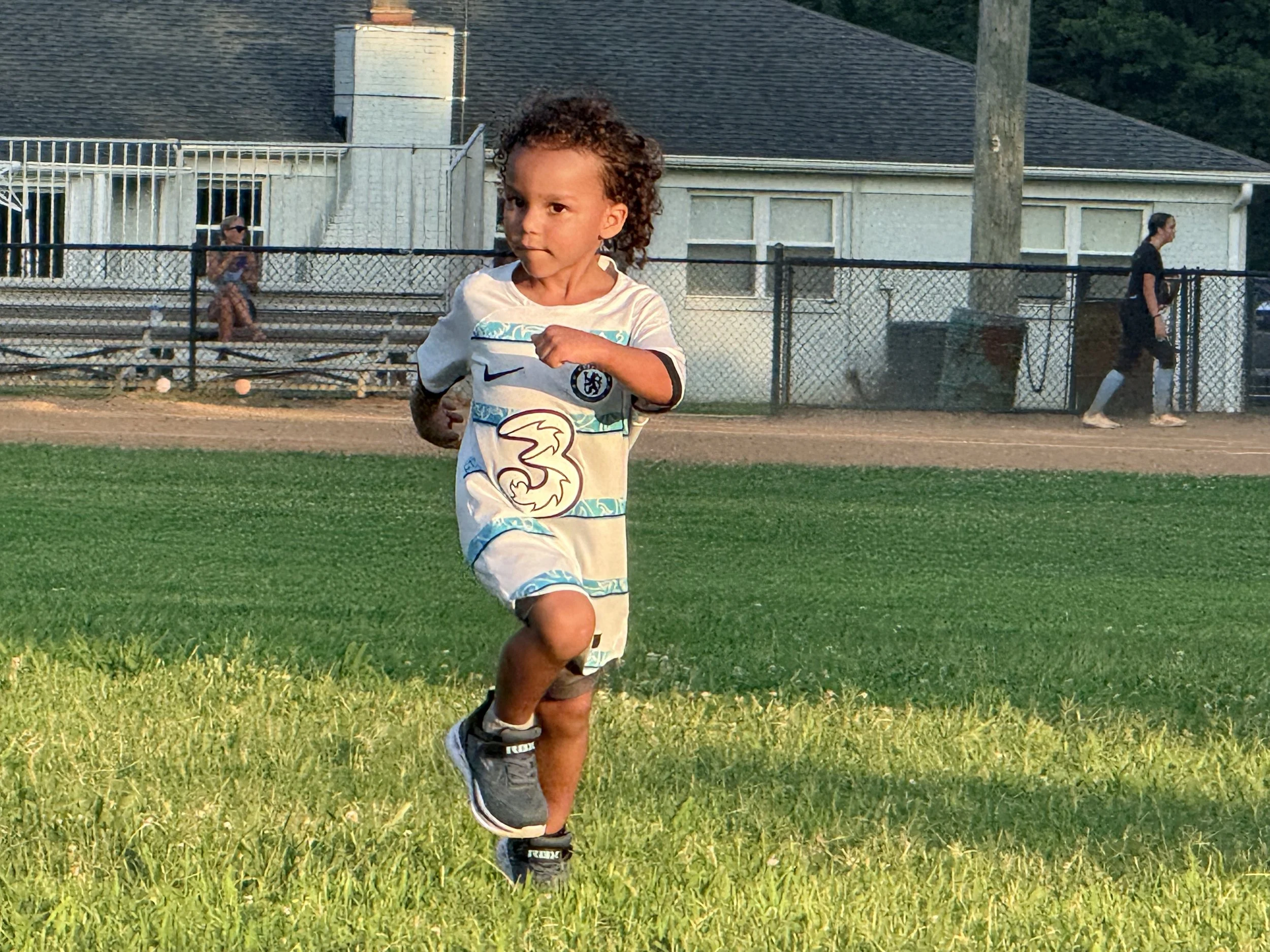 A young child running on a grassy field, wearing a white sports jersey with the number 3 and a Chelsea football club logo. In the background, there are a fence, a woman walking, and a white house with a chimney.