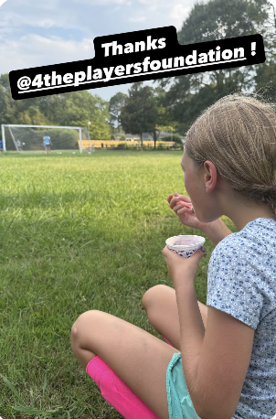 A young girl sitting on the grass at a soccer field, holding a snack and looking toward the field, with trees and a goalpost in the background, and a message thanking @4theplayerfoundation.