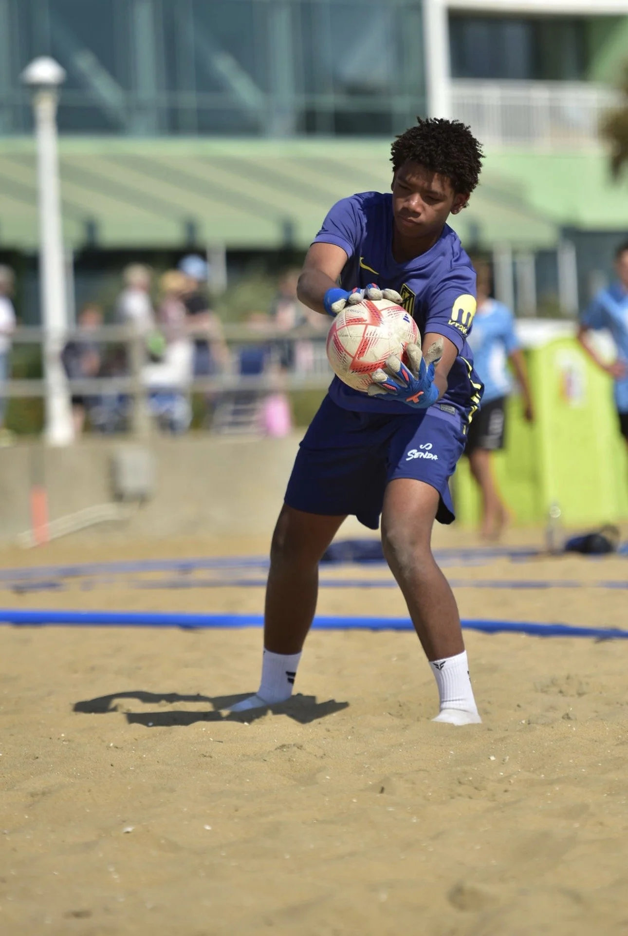 A young man wearing a blue sports uniform and gloves is practicing beach soccer goalkeeper drills on a sandy field during daytime. He is holding a soccer ball, preparing to throw or catch it, with a focused expression. There are blurred spectators and structures in the background.