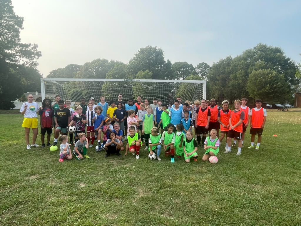Group of children and teenagers on a soccer field posing for a photo in front of a goal, some wearing green and red practice vests, with some holding soccer balls.