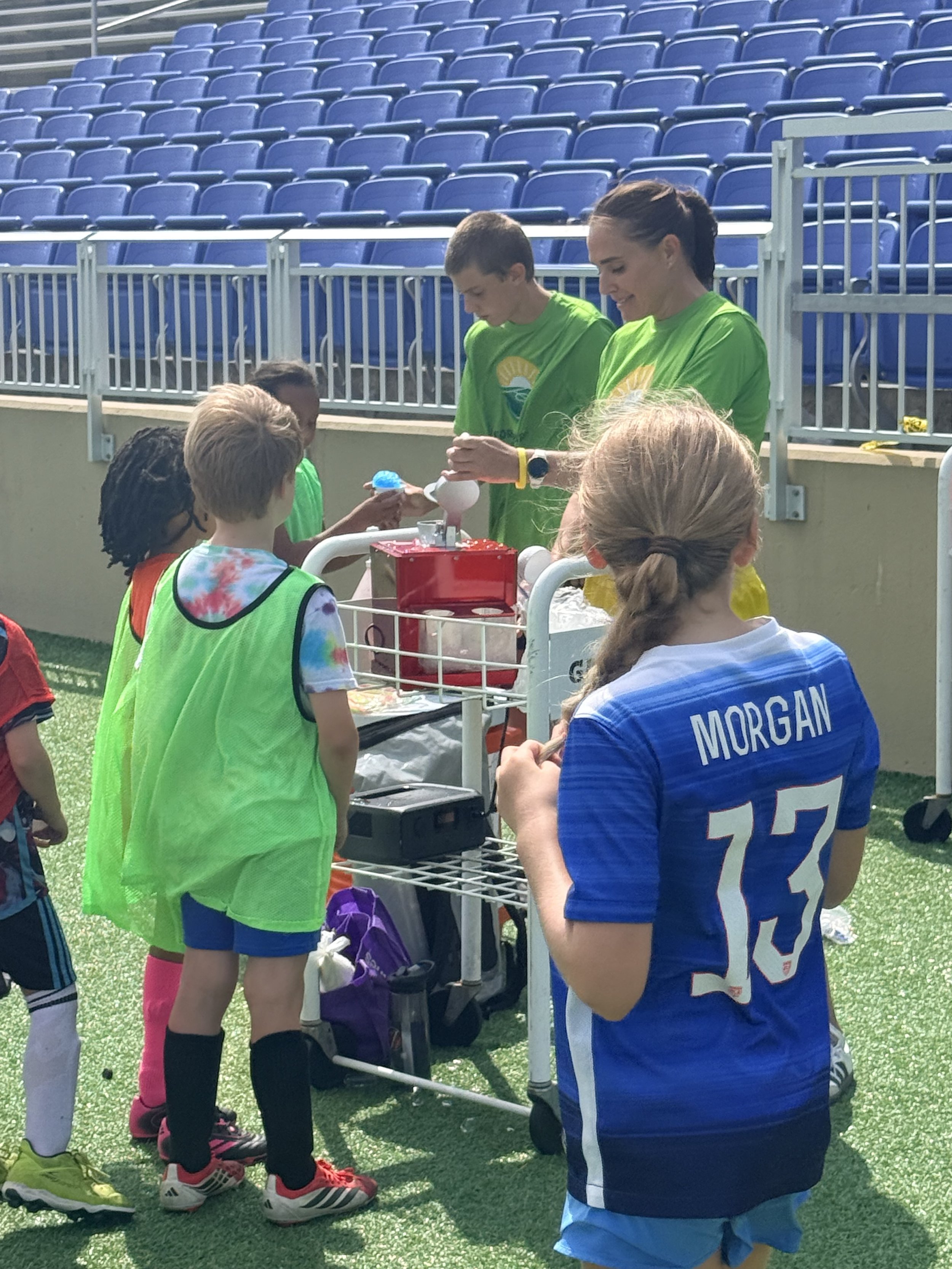 Football players and children at a hydration station on the field, some wearing neon green vests and others in blue and white jerseys, with blue stadium seats in the background.