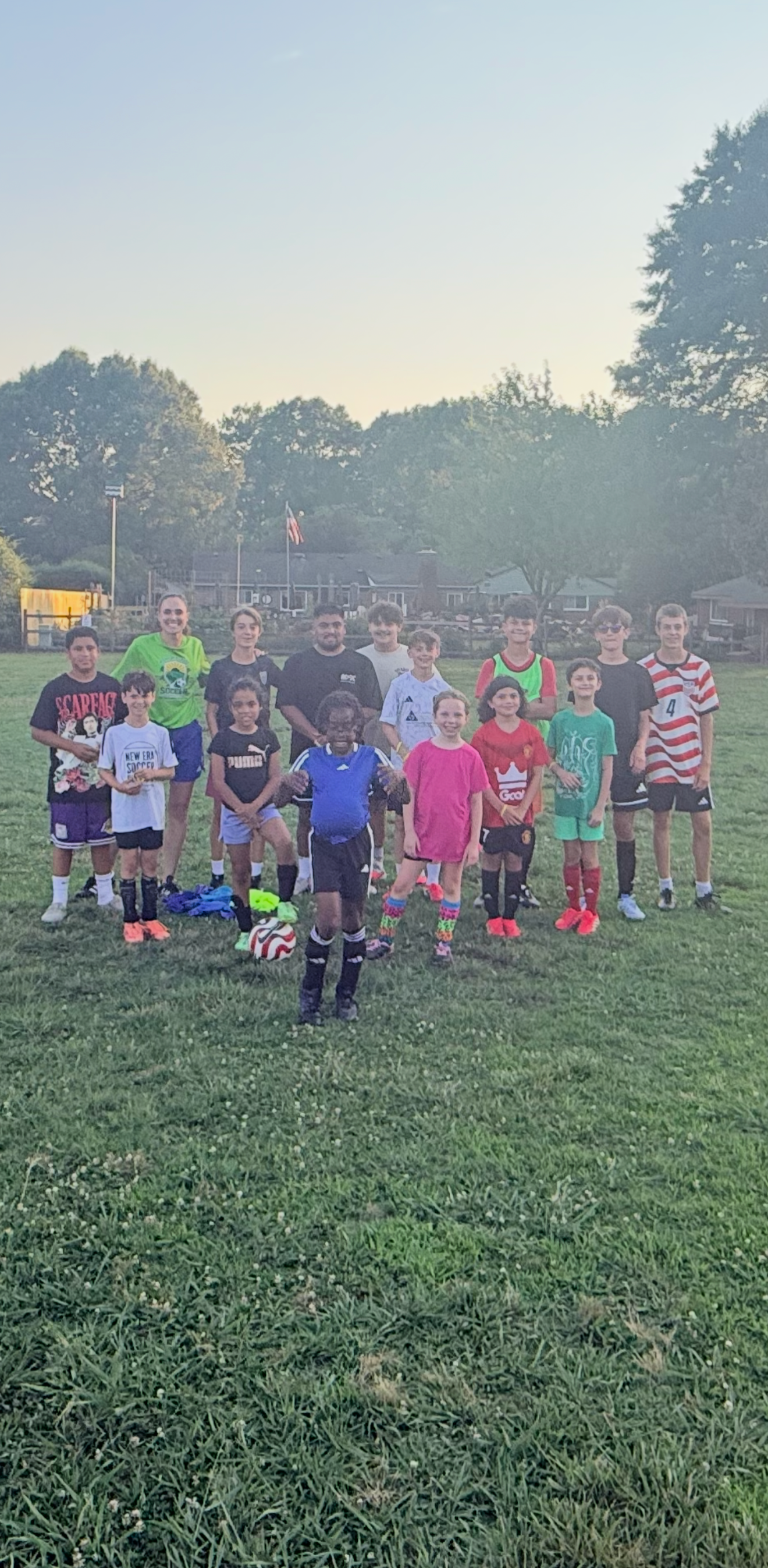 Group of children and adults on a grassy field, some wearing soccer jerseys, posing for a photo during sunset.