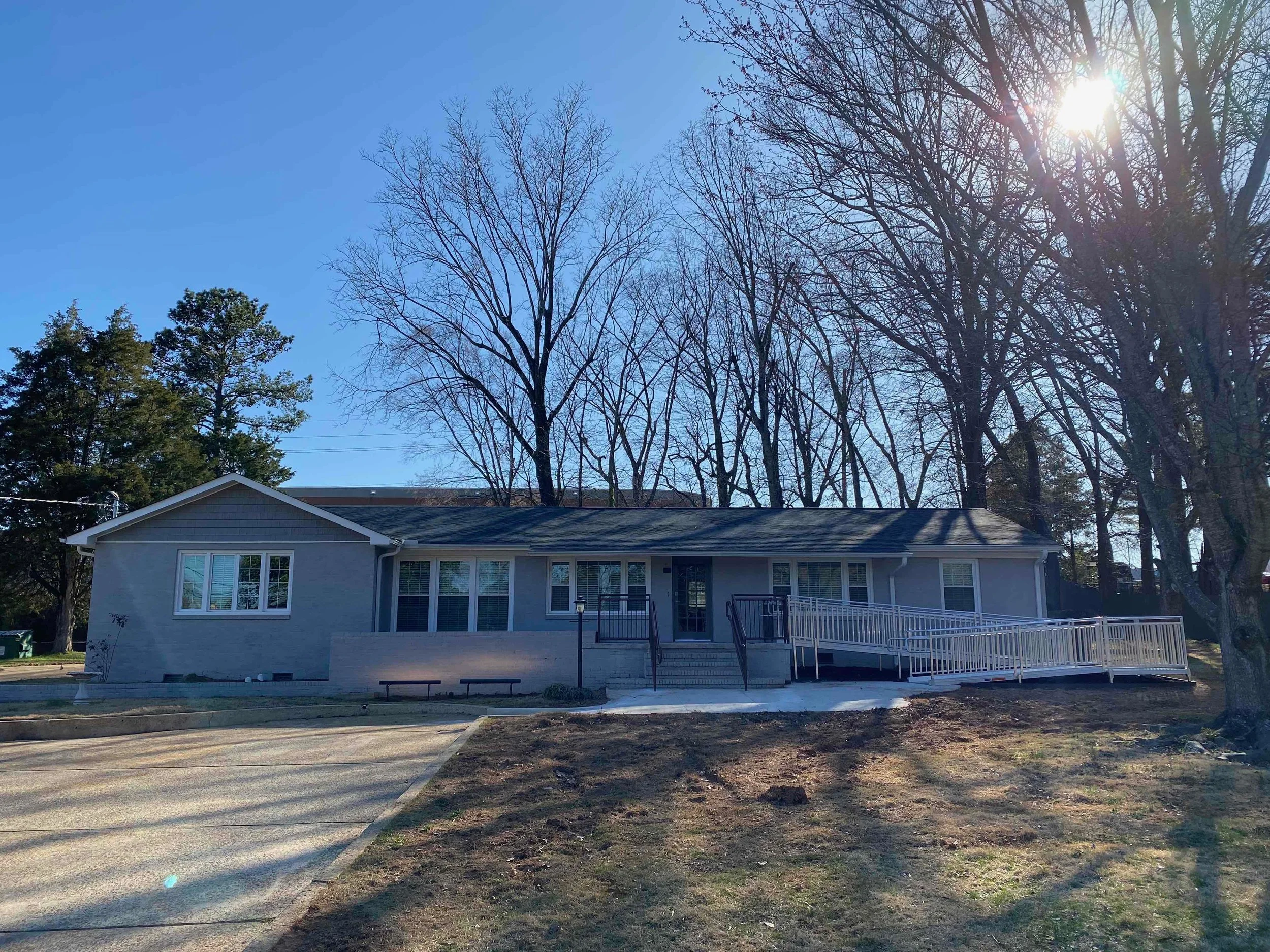 A single-story house with grey siding, white trim, and a black roof, with a ramp leading up to the front door. The yard is partly bare with patches of grass, and large leafless trees are behind the house with the sun shining through.