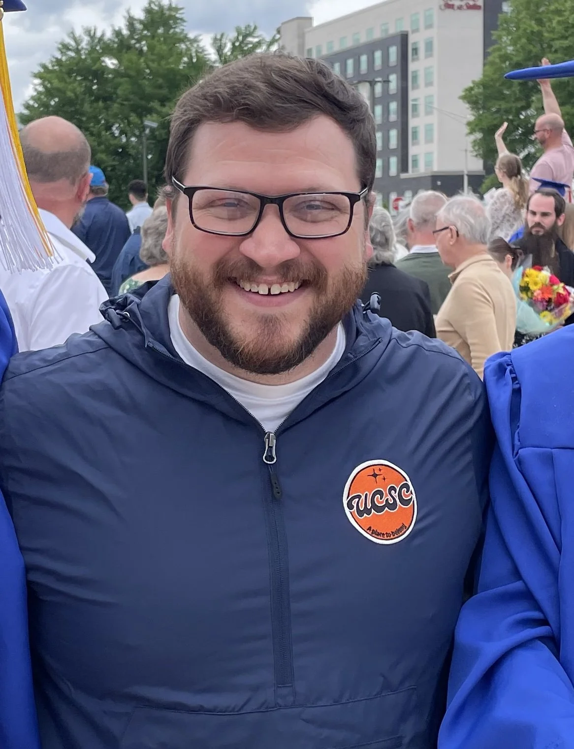 A smiling man wearing glasses and a navy blue jacket with a NASA logo on it. He is at a graduation ceremony with many people, some holding flowers, in an outdoor urban setting.