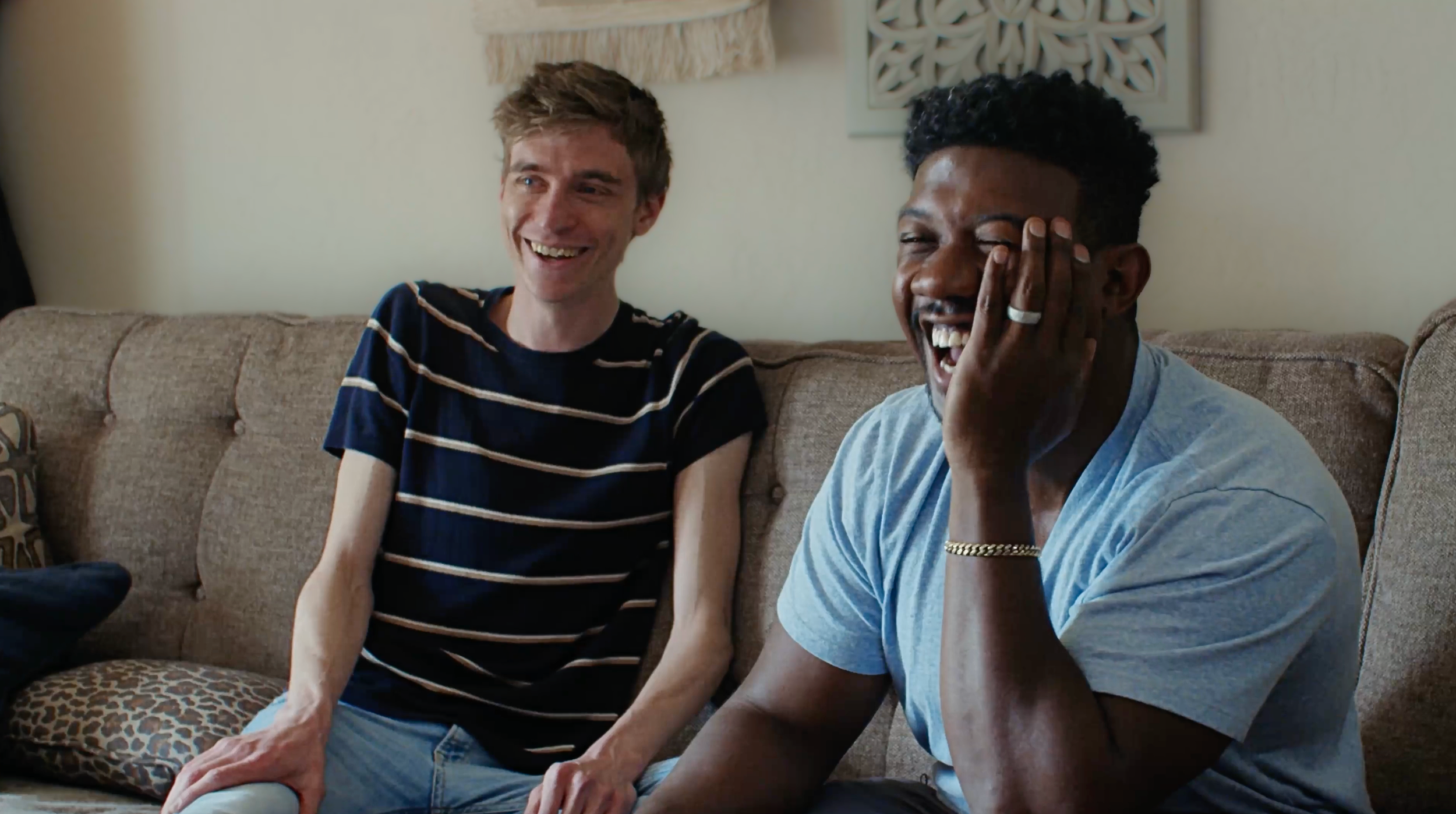 Two men sitting on a beige couch, laughing and smiling. One is wearing a striped navy and white t-shirt, and the other is in a light blue shirt with a gold bracelet and ring.