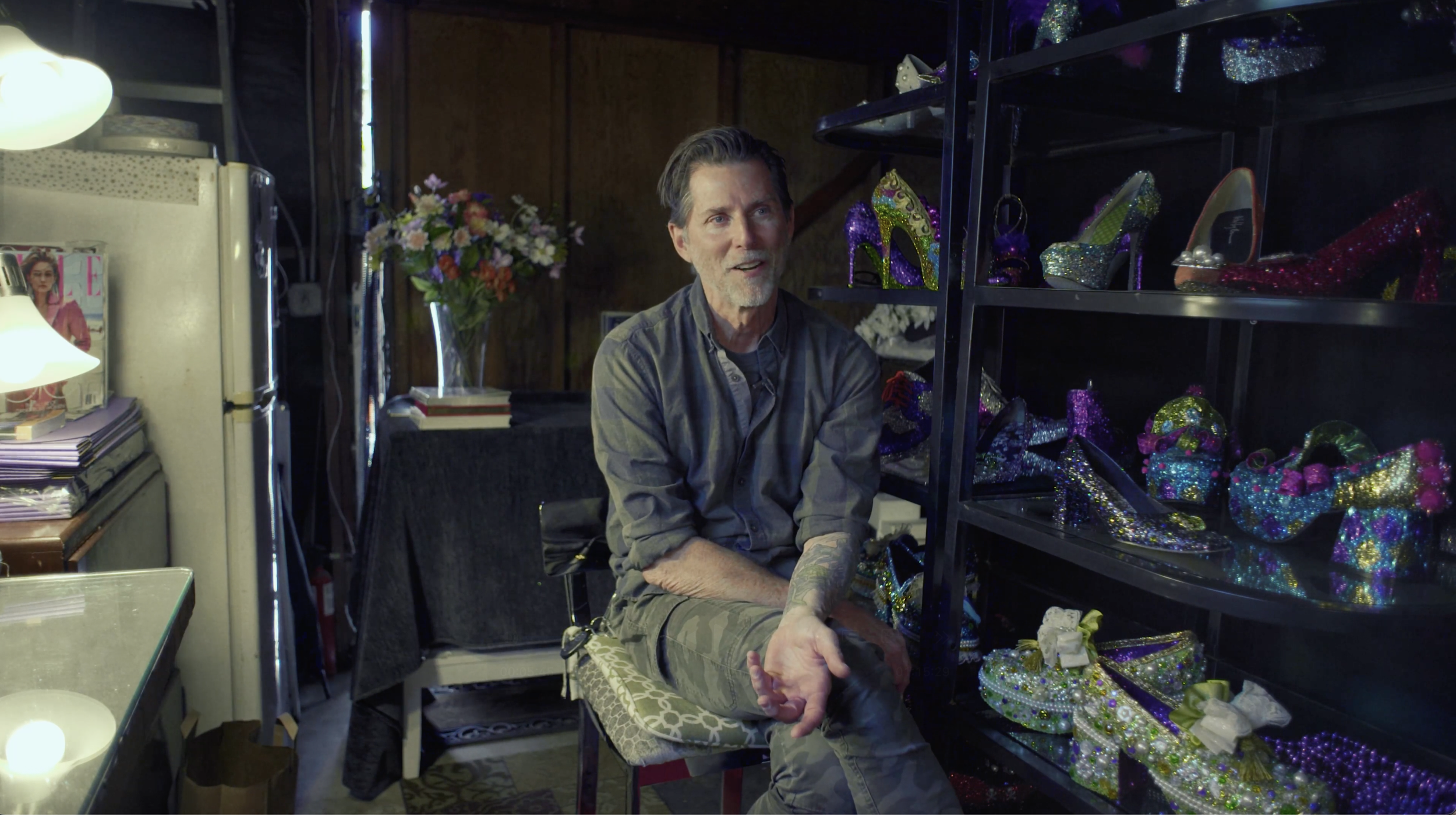A man with gray hair, beard and tattoos sitting on a bench in a room filled with colorful, glittery high-heeled shoes on shelves.