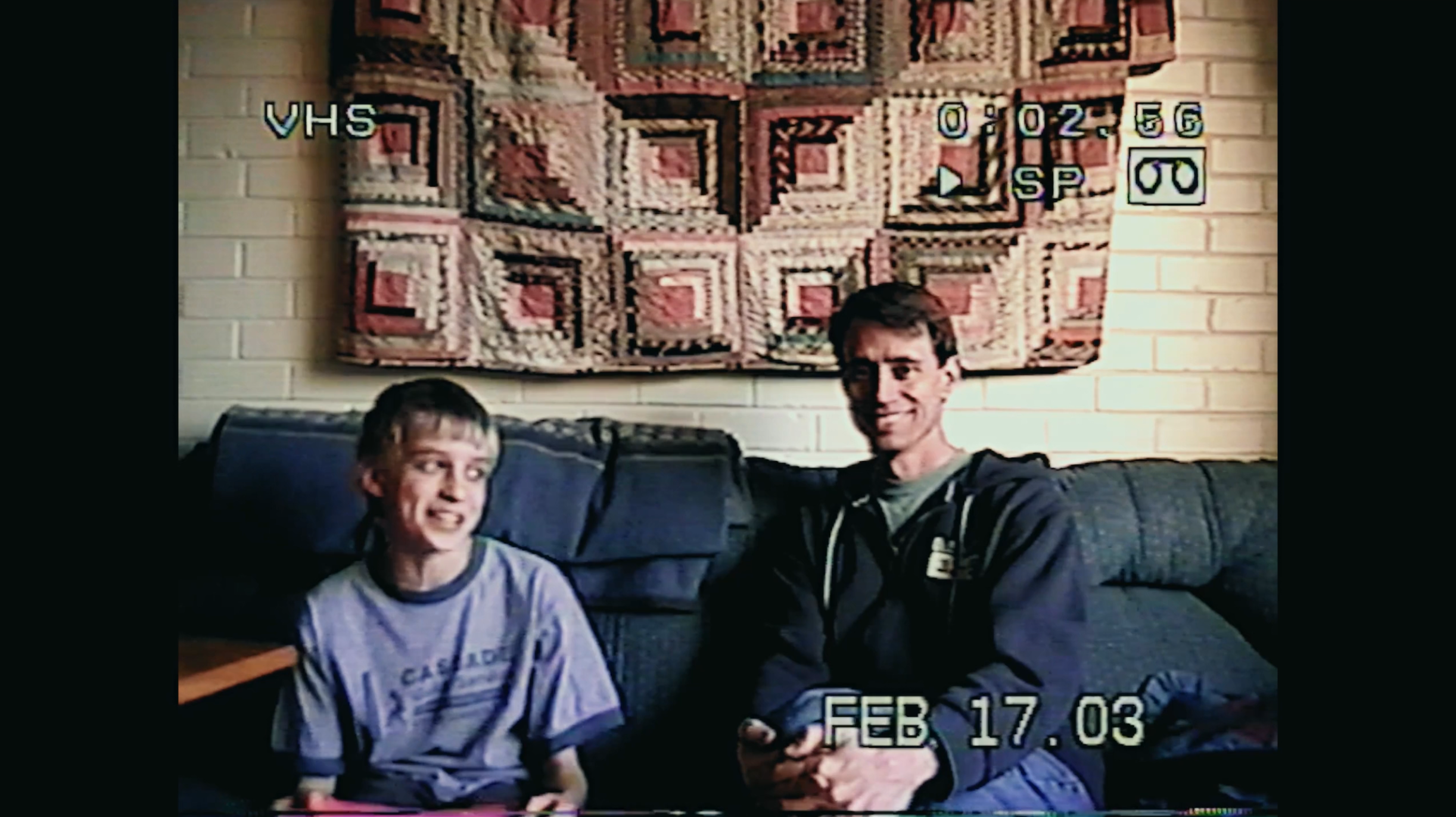 A young boy and an adult man sitting on a sofa in a living room, smiling at the camera. Behind them is a quilt hanging on a brick wall. The image has a timestamp indicating February 17, 2003, and VHS recording details.