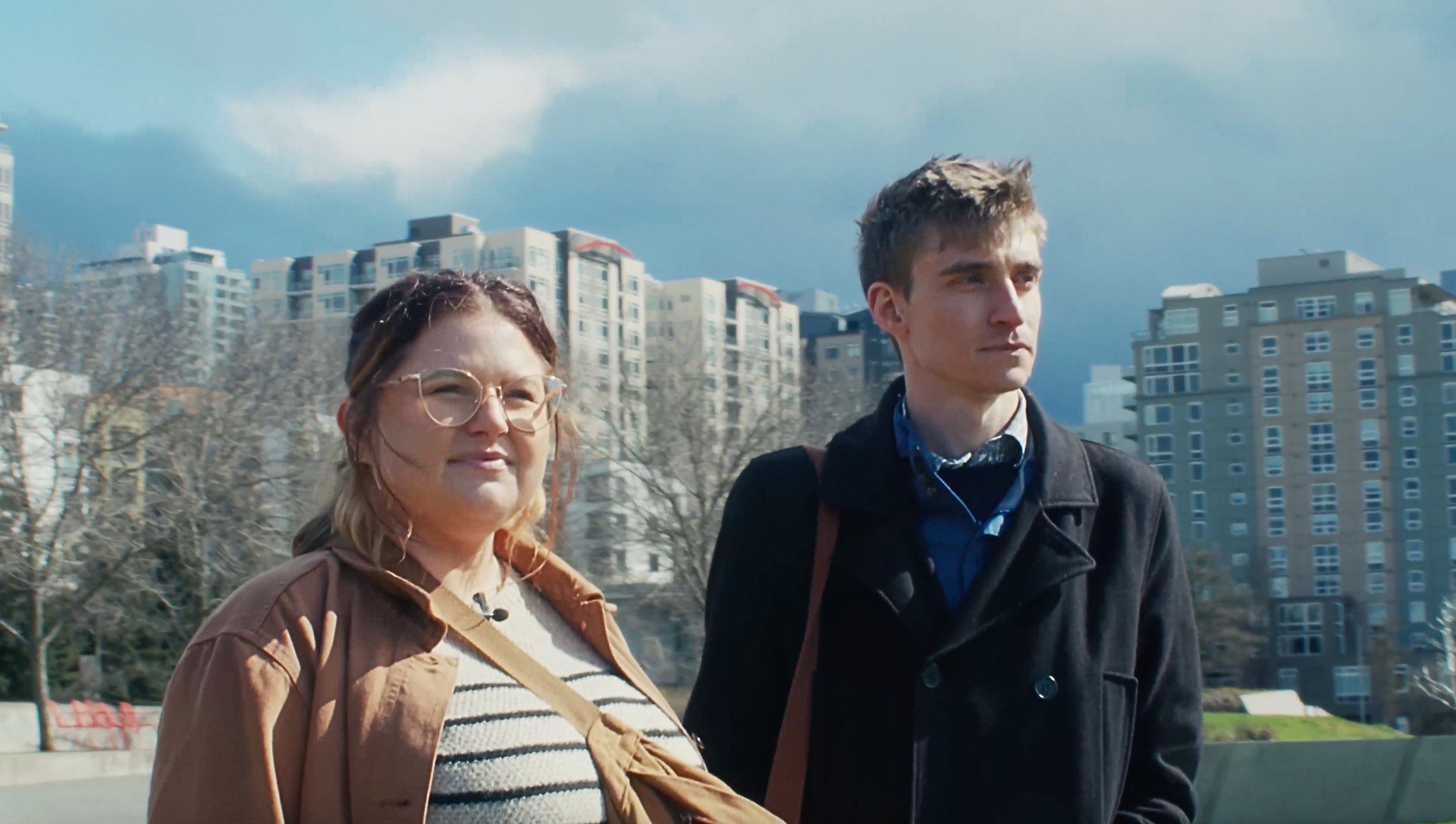 A woman with glasses and a man with short hair standing outdoors with a cityscape of high-rise buildings behind them, under a cloudy sky.