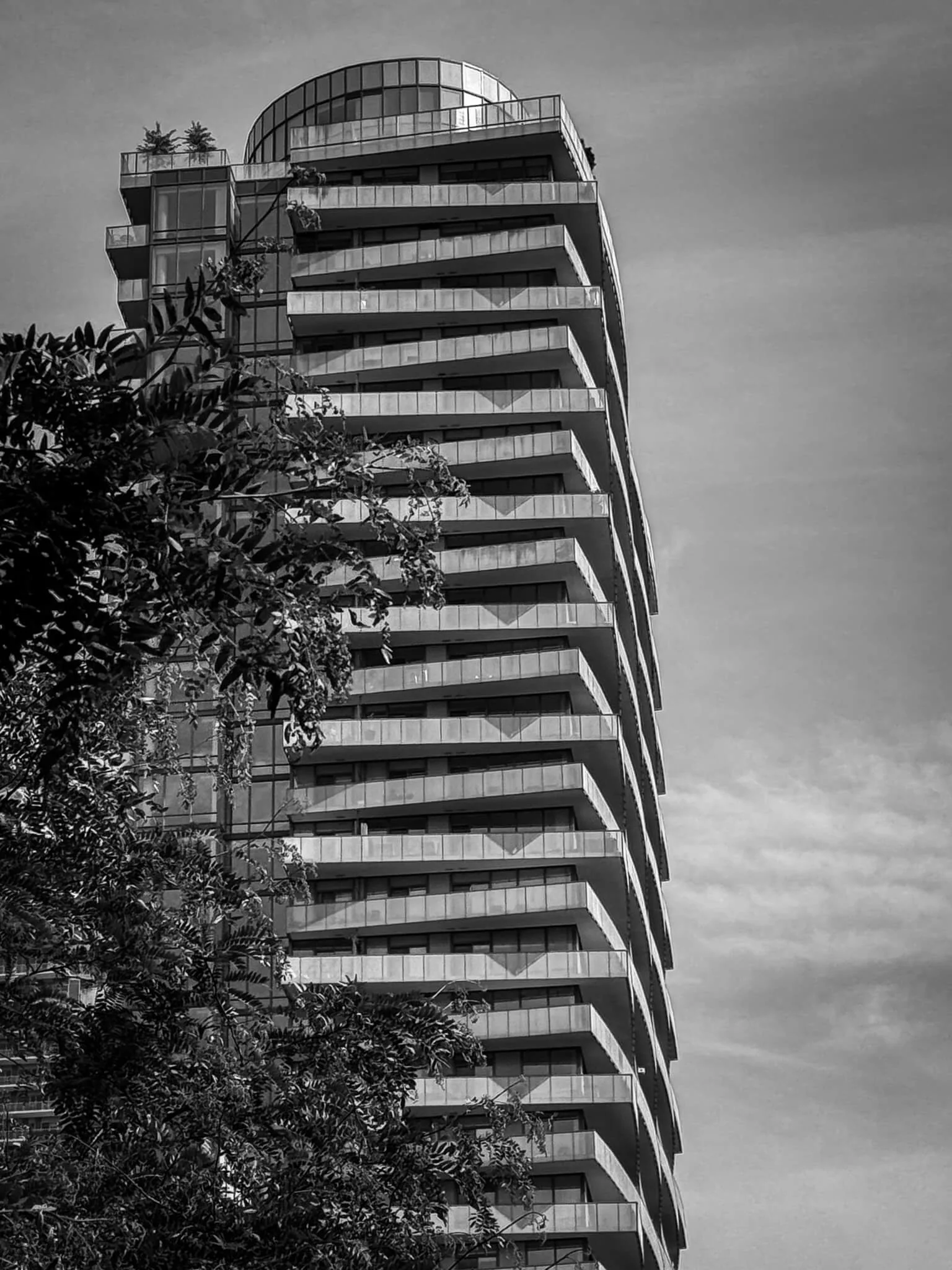 A modern high-rise building with multiple balconies, glass windows, and rounded architecture at the top. There are trees in the foreground and a partly cloudy sky in the background.
