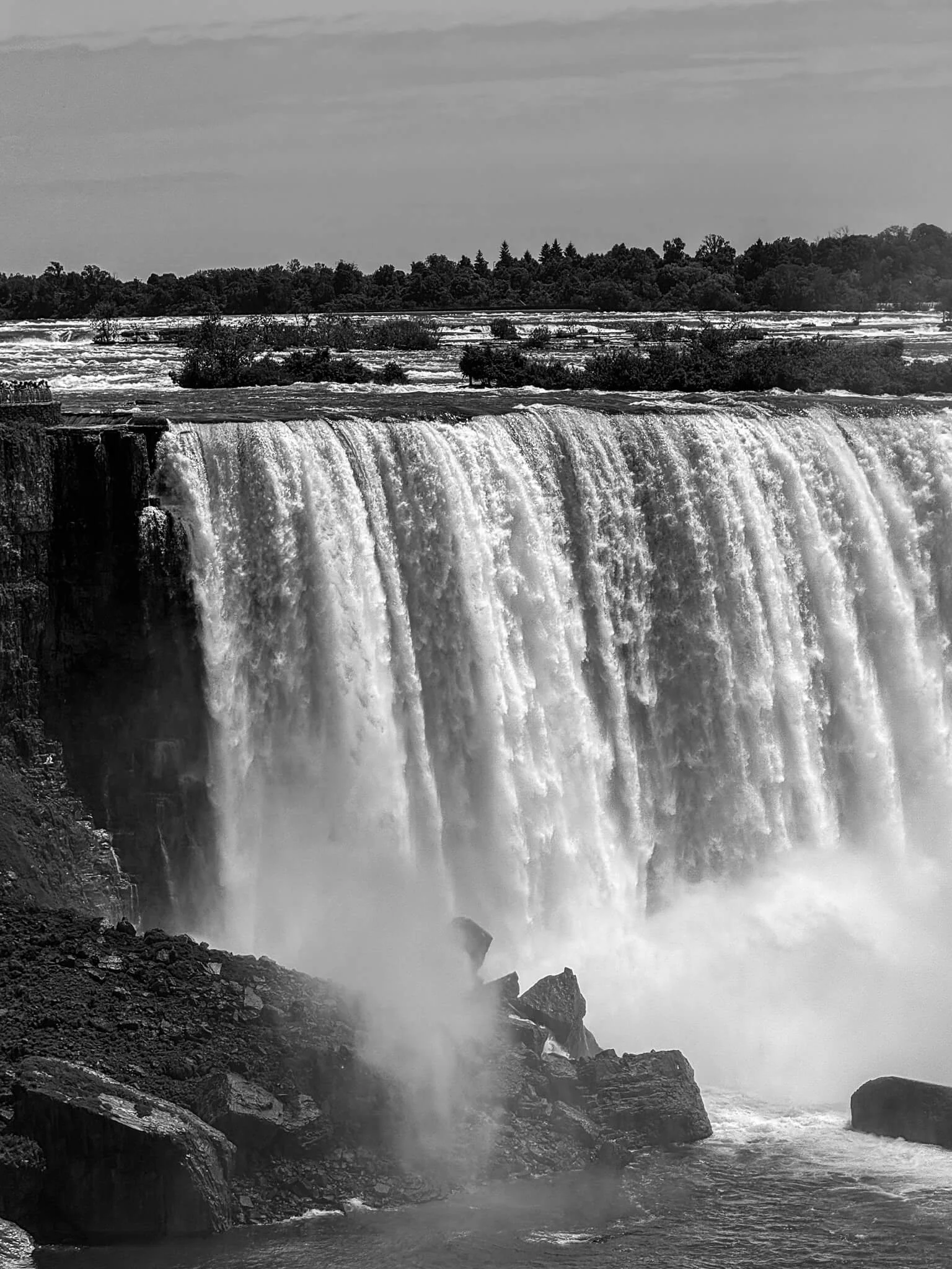A waterfall cascading over a cliff with rocks and mist at the bottom, and trees and a river in the background.