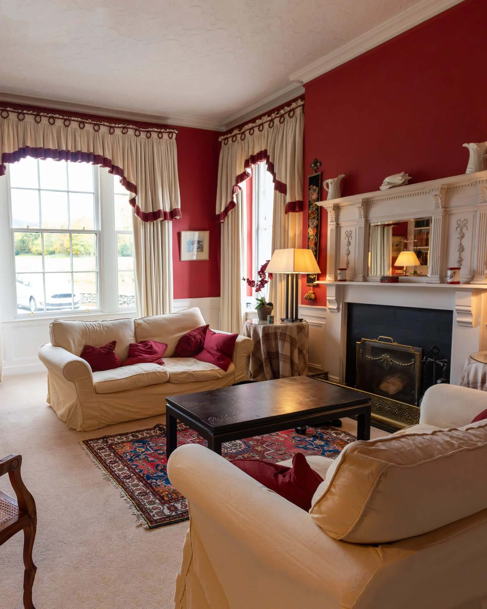 Living room with beige sofas, a black coffee table, a rug, a fireplace with a mirror above it, cream curtains with red trim, a side table with a lamp and vase, and red walls.