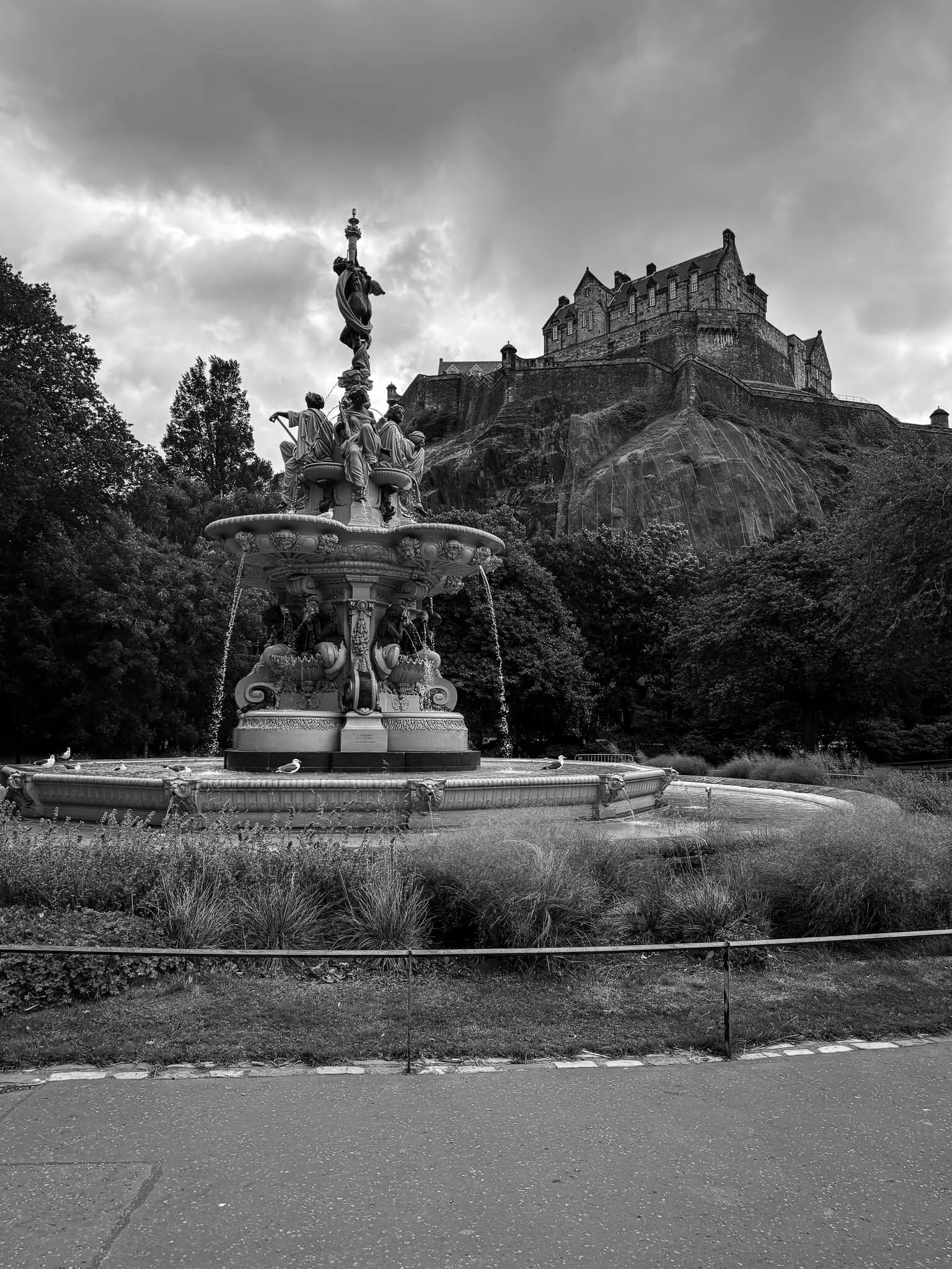 A black and white photograph of a decorative fountain with statues on top, surrounded by grass and trees, with a large castle on a hill in the background and cloudy sky overhead.