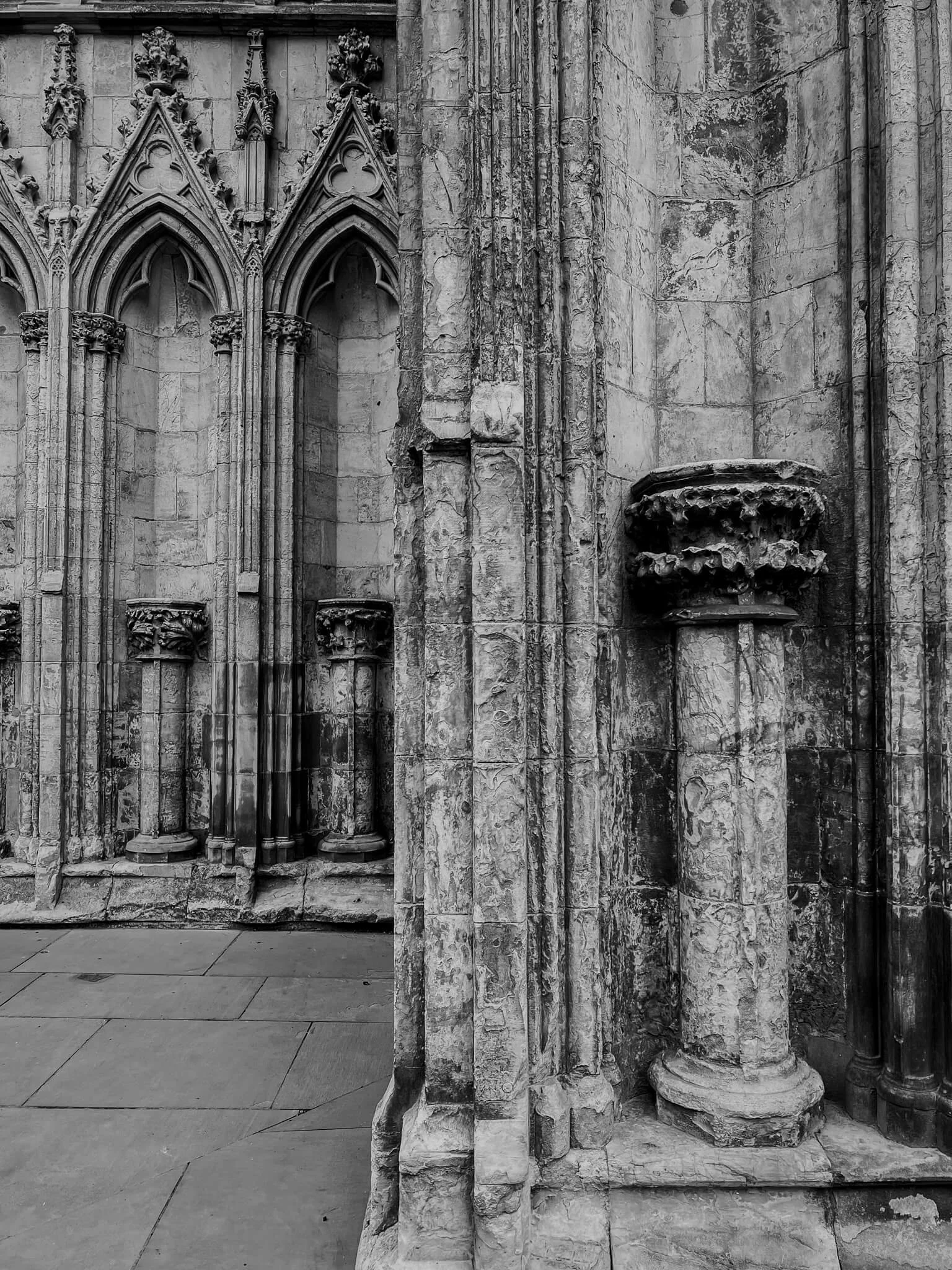 Black and white photo of a stone gothic cathedral wall with ornate arches, columns, and a weathered column base.