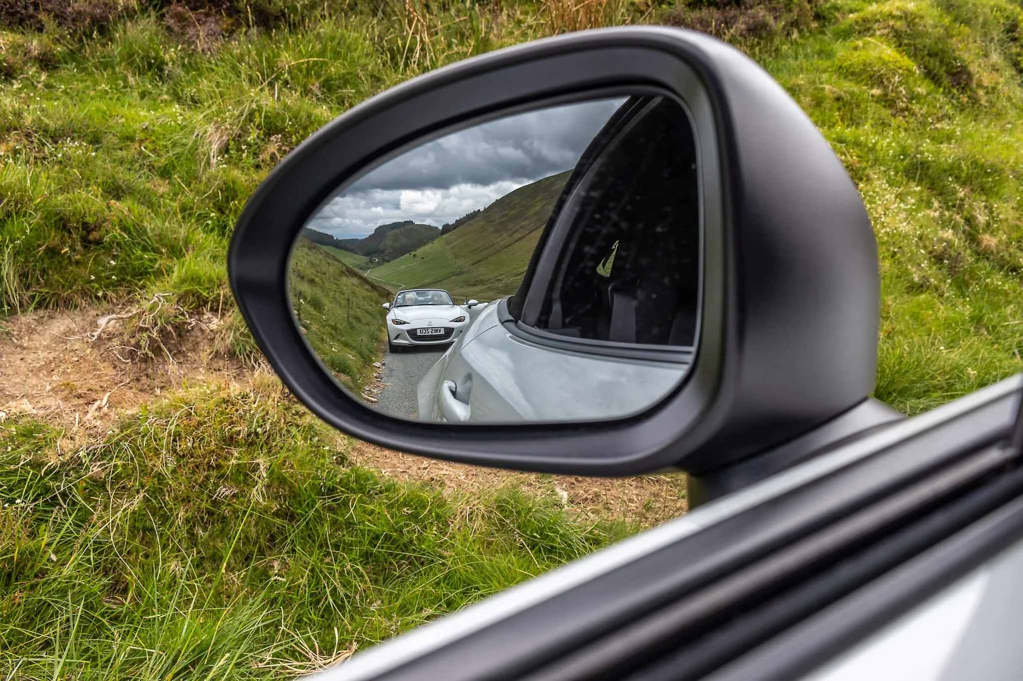View of a white sports car on a narrow mountain road, seen through the side mirror of another car, with grassy hills and a cloudy sky in the background.