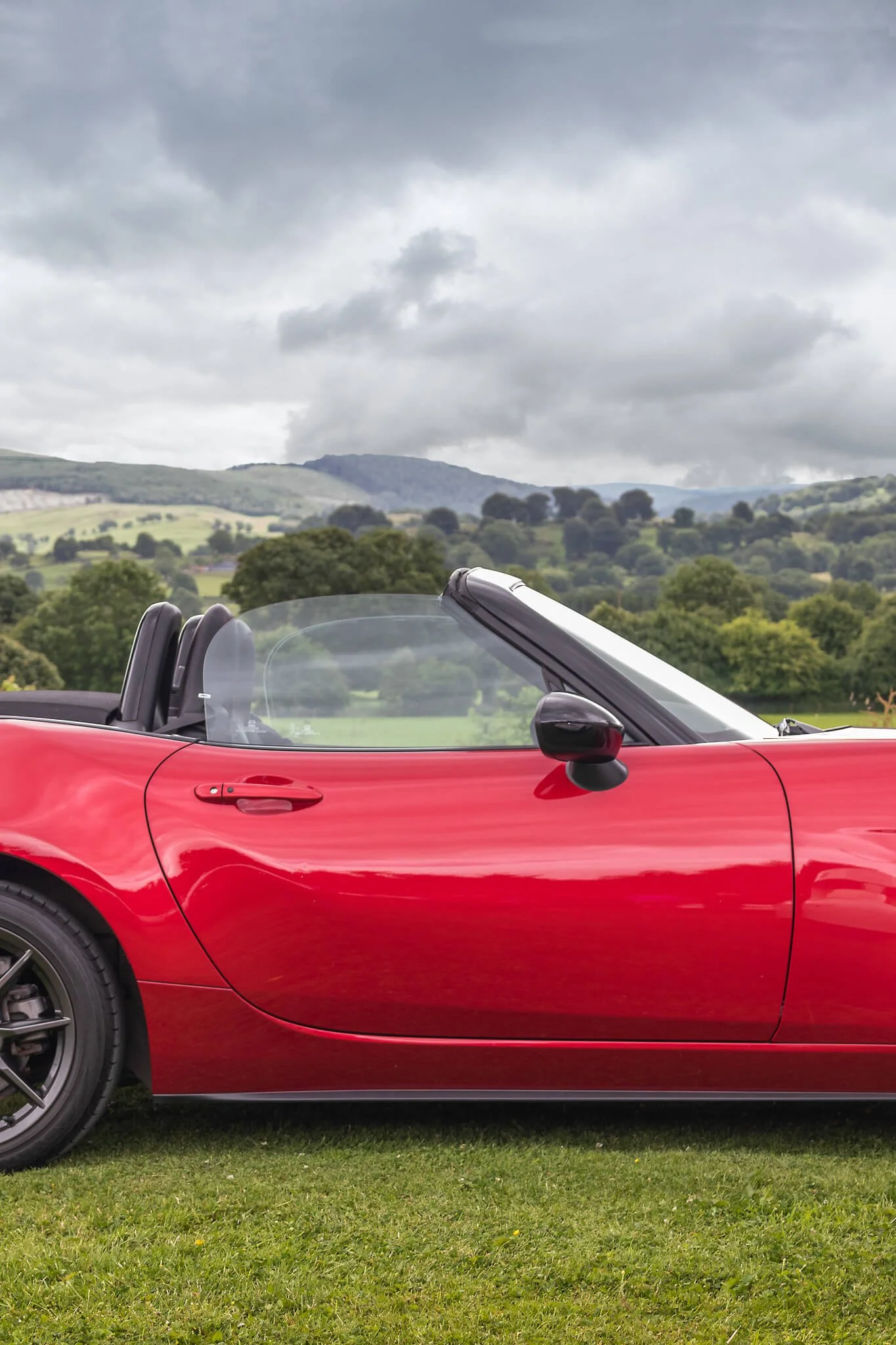 A red convertible sports car parked on grass with a scenic countryside landscape and cloudy sky in the background.